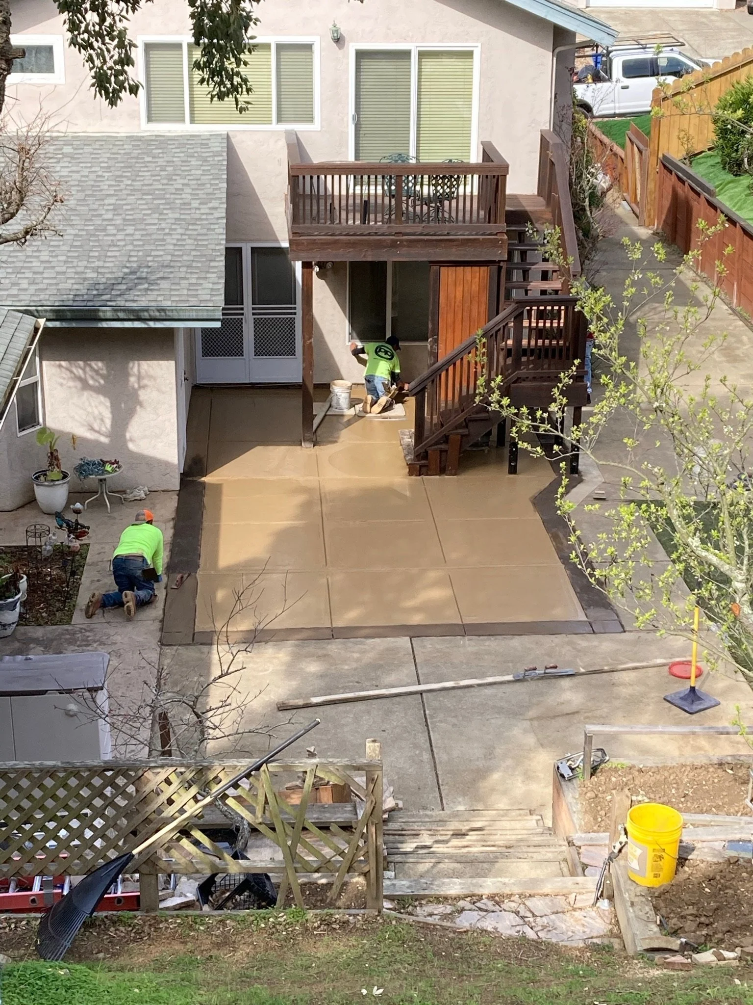 Two workers in bright green shirts are working on a home improvement project on the backyard patio, installing a concrete slab. The patio is partially fenced with a wooden railing and stairs leading to the yard and the house. There's a yellow bucket 