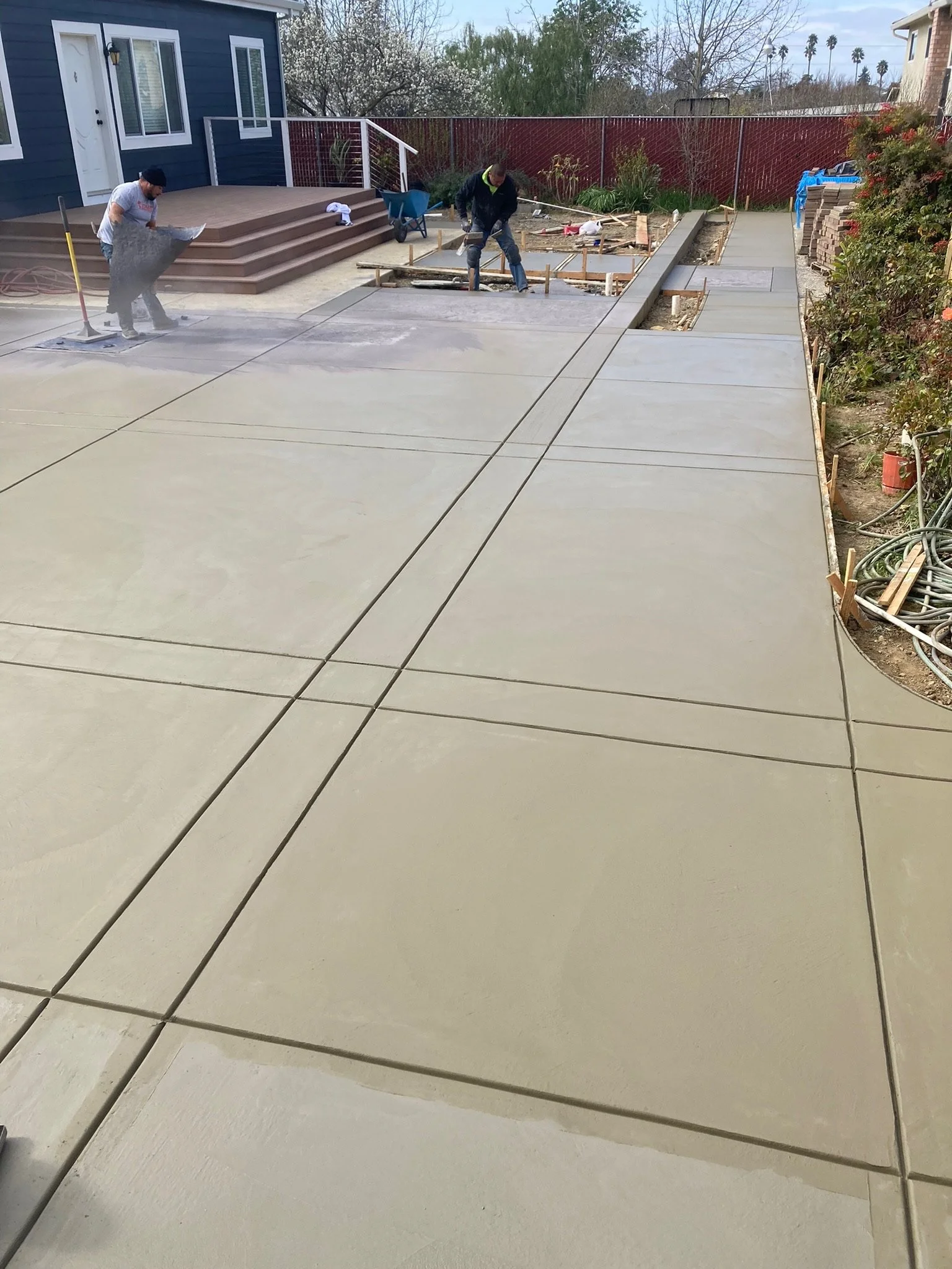 Two construction workers are laying concrete on a driveway in a backyard. One worker is spreading concrete with a tool, while the other is working with a concrete saw. There is a staircase leading to a house with blue walls on the left side of the im