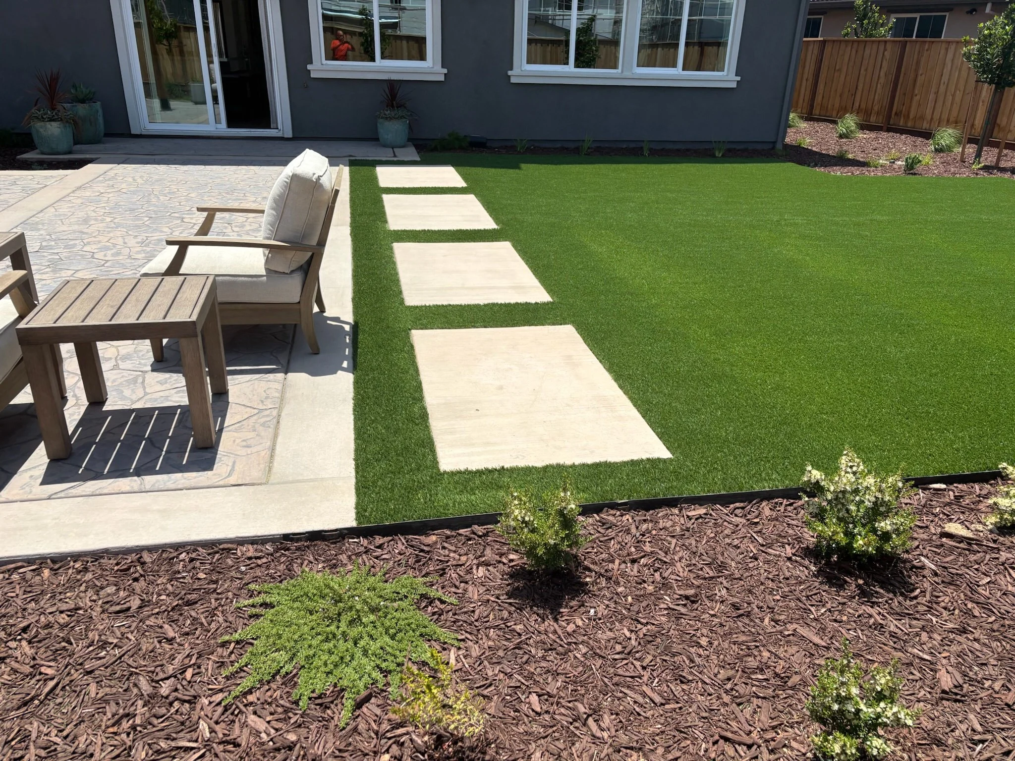 A backyard patio with a small grassy area, concrete stepping stones, a wooden chair and table, potted plants, and a landscaped flower bed with mulch.
