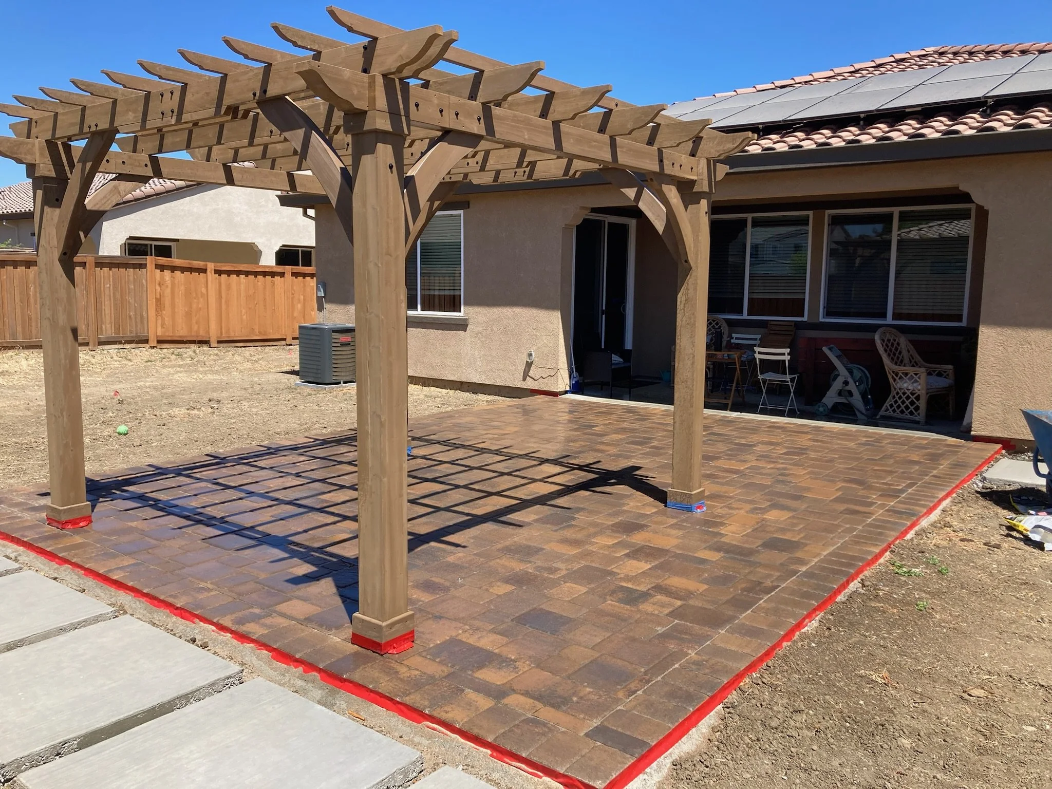A backyard with a new wooden pergola under construction, paved patio, adjacent house with beige stucco walls, windows, and solar panels on the roof, and a wooden fence in the background.