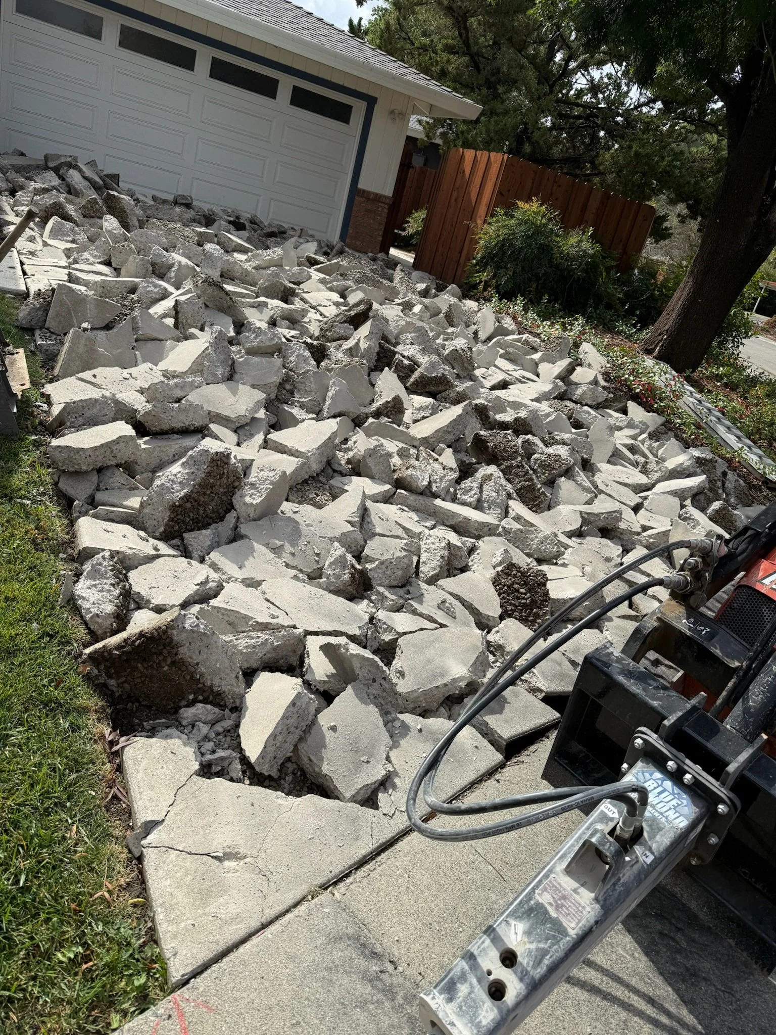 A yard undergoing excavation with broken concrete slabs and debris, a tree providing shade, a wooden fence, and construction equipment visible.