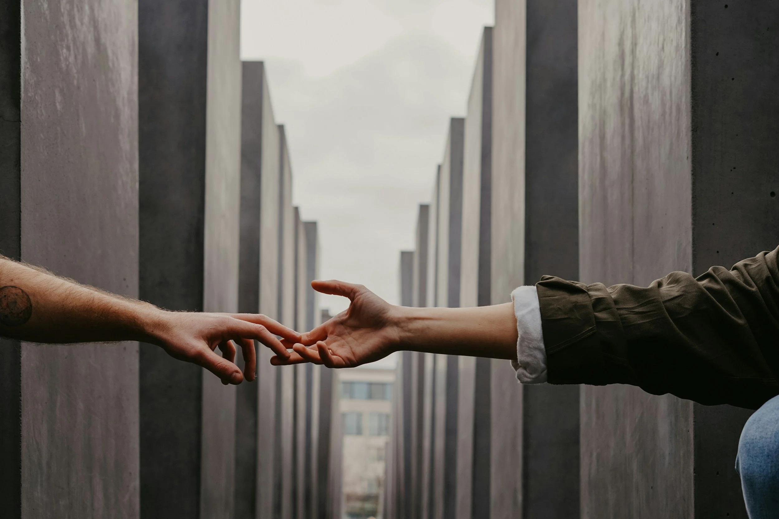Two people reaching out and touching hands in the middle, with a background of tall concrete structures and cloudy sky.