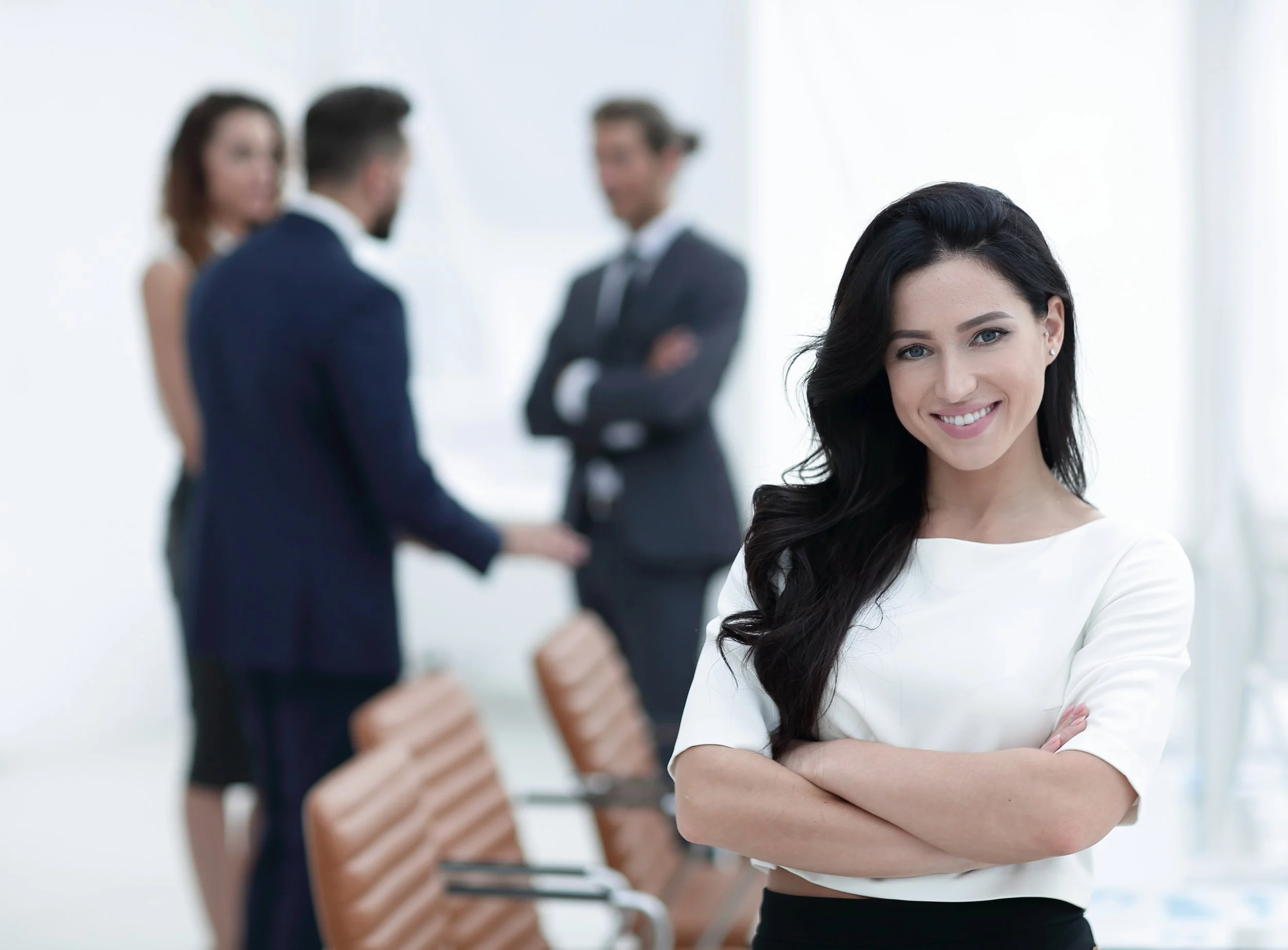 A woman with black hair and blue eyes standing with her arms crossed, smiling at the camera, with three out-of-focus people in business attire in the background in a professional setting.