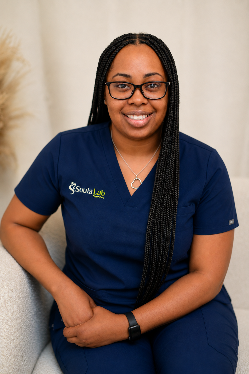 A woman in a dark blue scrubs uniform with 'Soula Lab Services' embroidered on it, sitting on a light-colored sofa, smiling at the camera, wearing glasses and a necklace with a heart-shaped pendant.