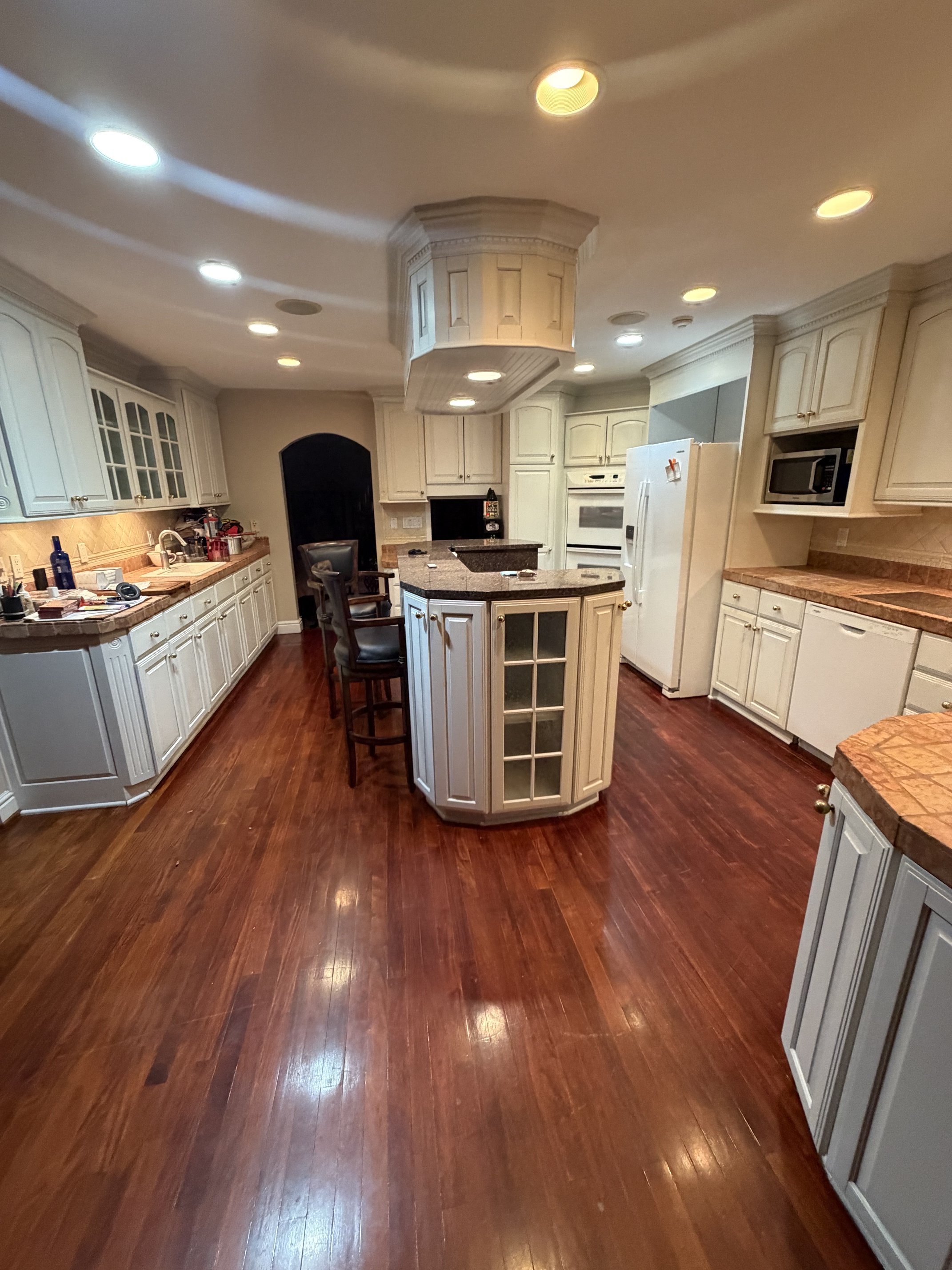 A spacious kitchen with white cabinets, a central island with a granite countertop, hardwood flooring, and recessed ceiling lights.