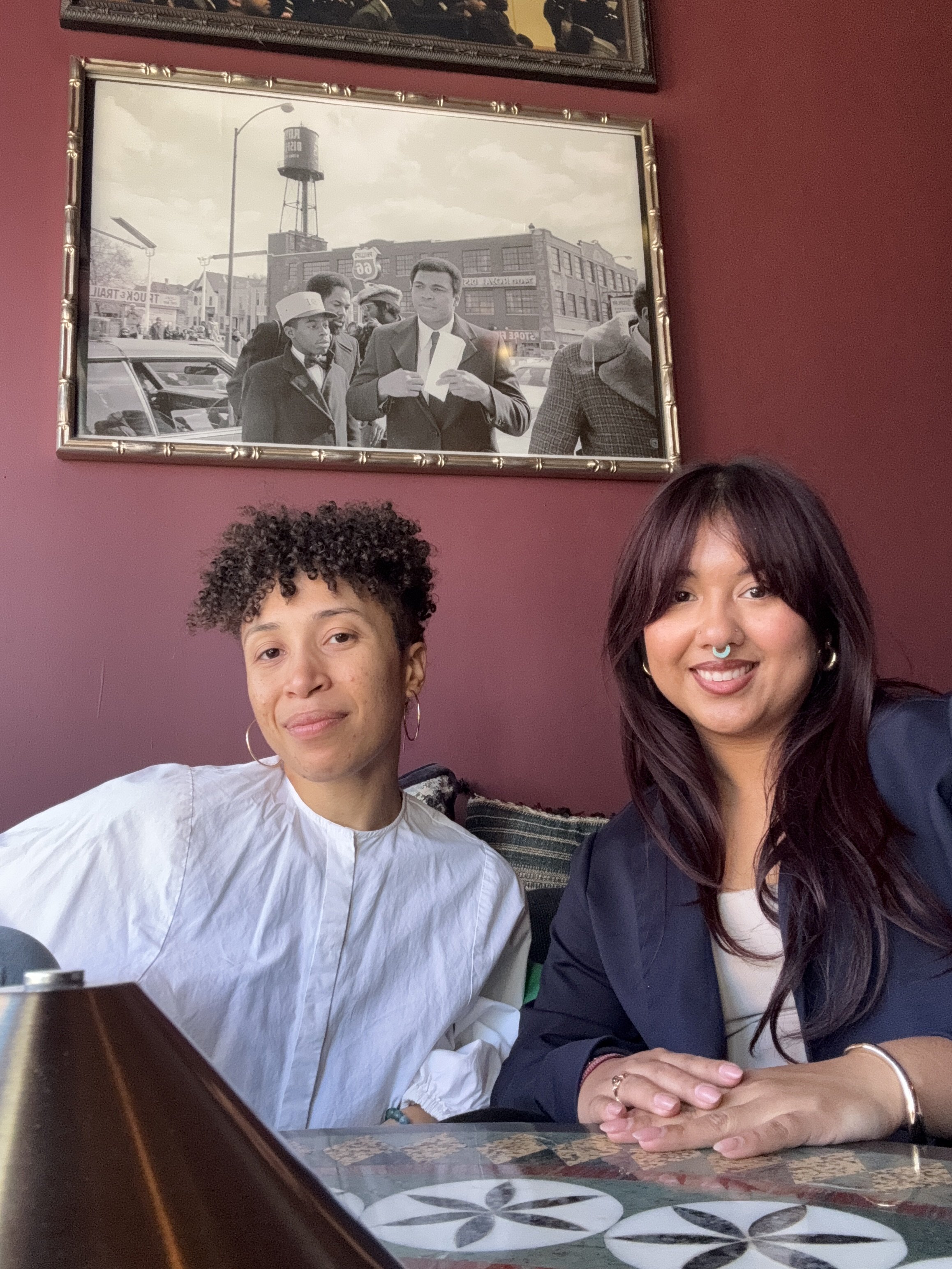 Two women sitting at a table in a restaurant. Behind them, there is a framed black and white photo on the wall showing a group of men, including one man holding a piece of paper. The restaurant has a maroon-colored wall and a decorative tile table.