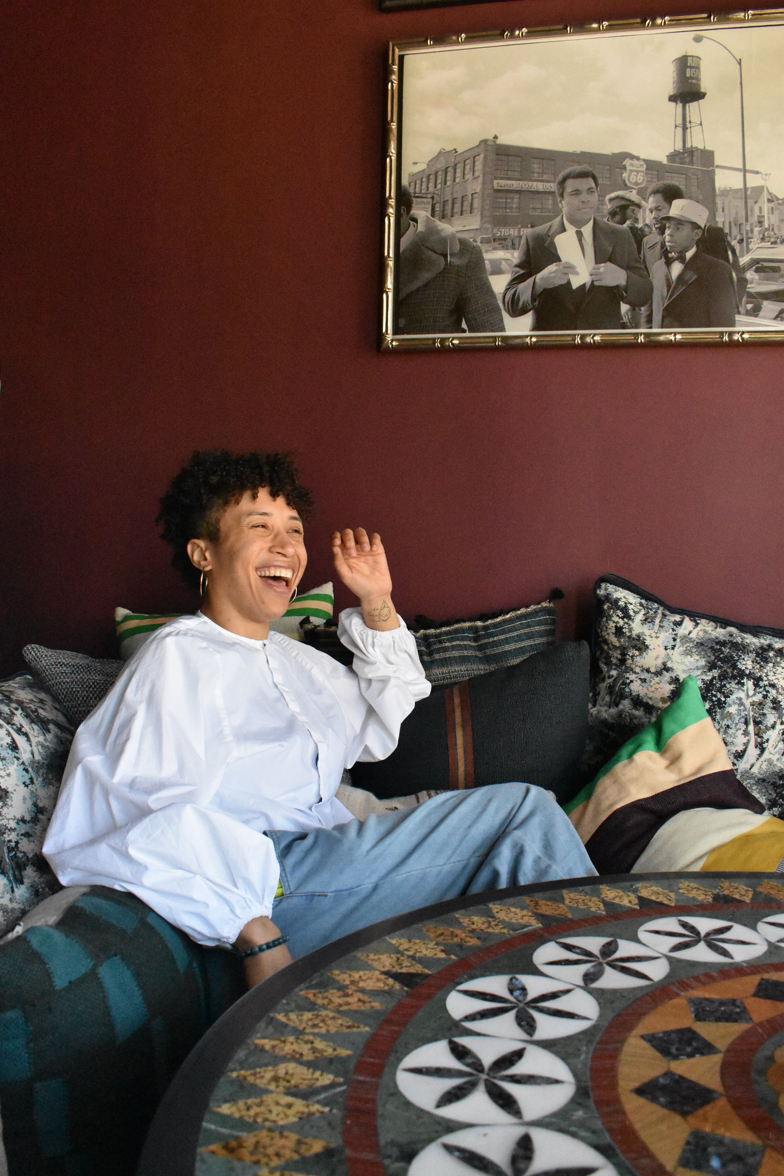 A woman with short curly hair, wearing a white shirt, sitting on a couch with patterned pillows, laughing and waving. Behind her, there's a wall with a framed black-and-white photograph of a historical street scene.