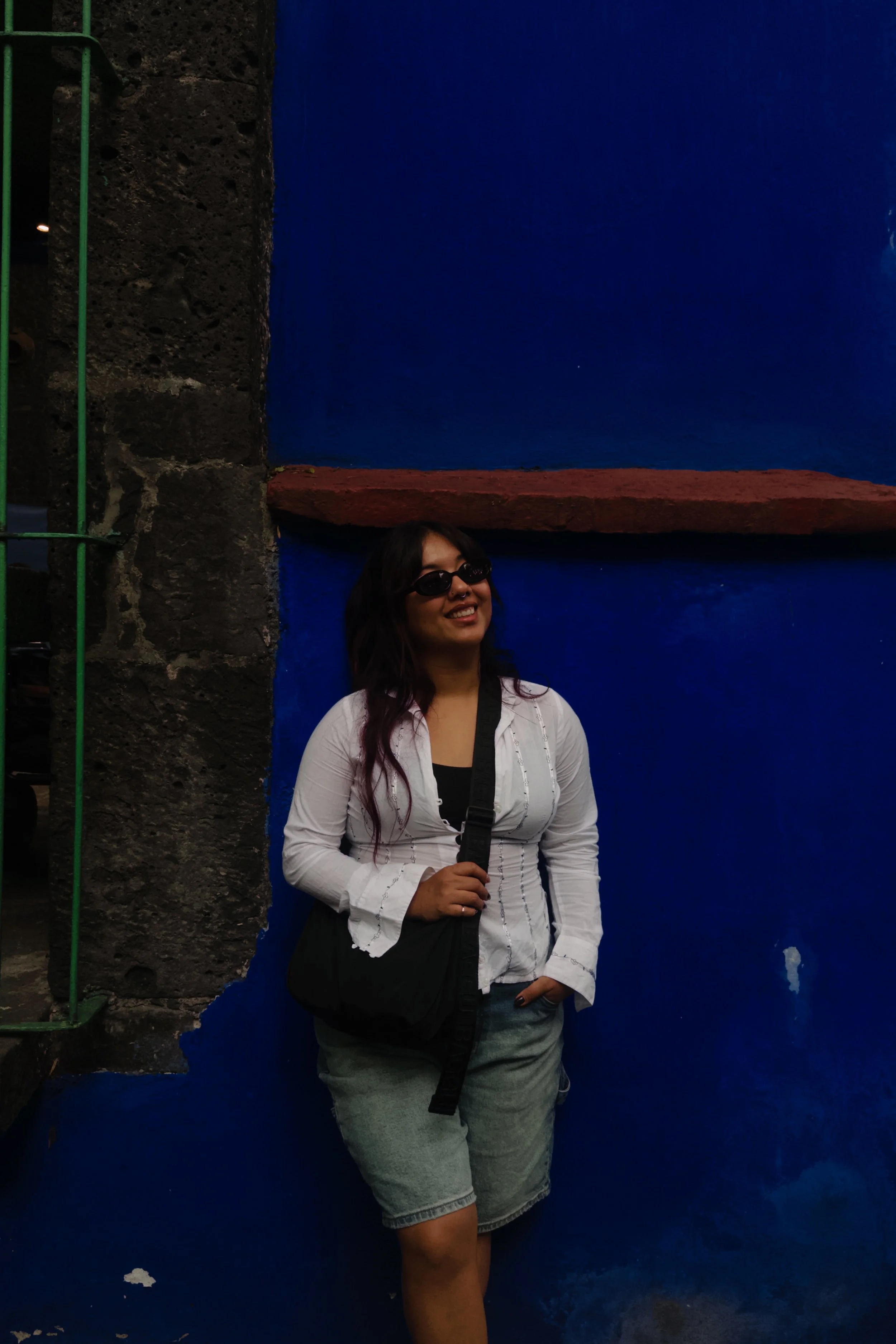 A young woman with long dark hair, wearing sunglasses, a white jacket, and gray shorts, is standing against a blue wall, smiling, while holding a black strap in her right hand.