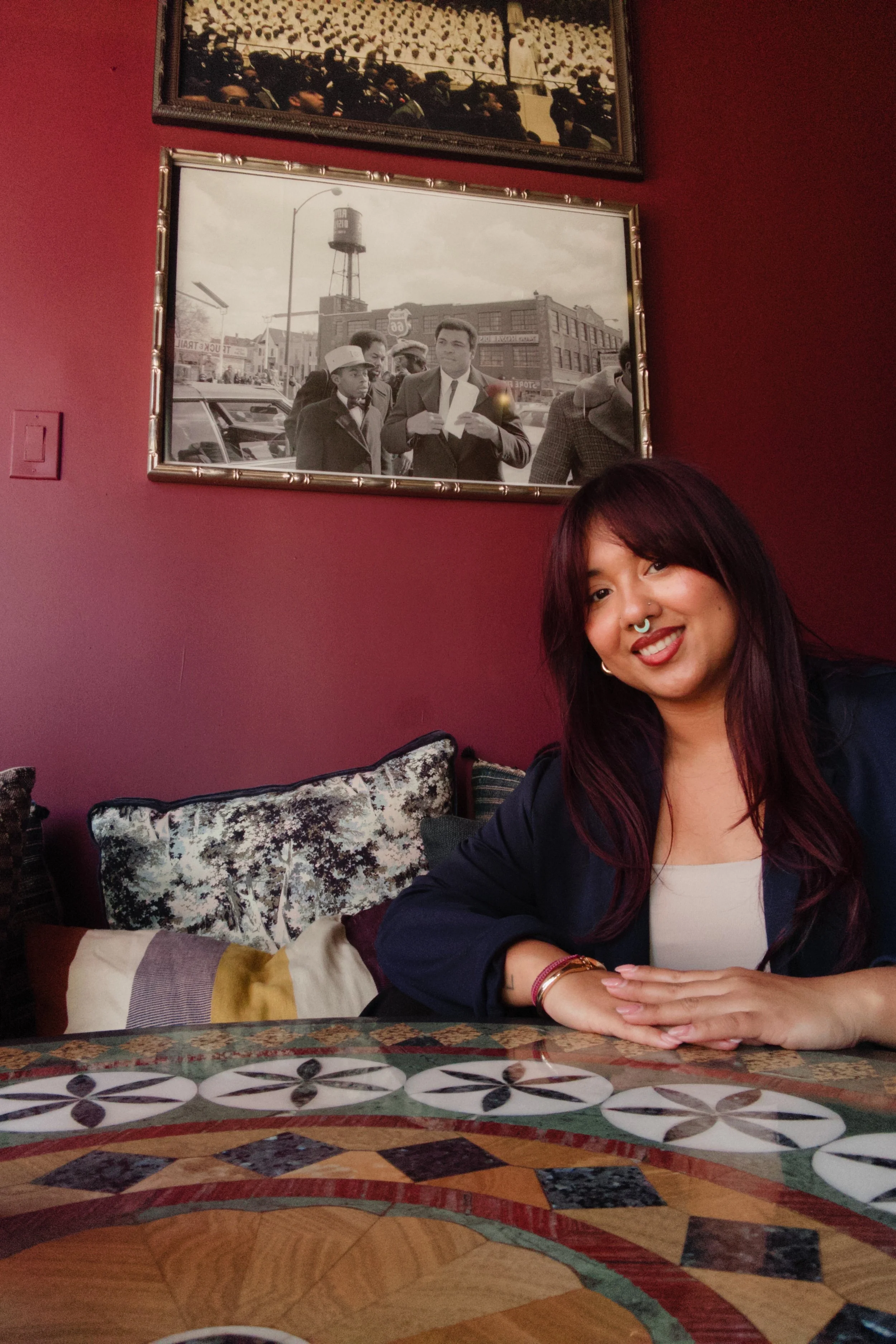 A woman with dark hair and a septum piercing smiling at a restaurant table with patterned cushions and decor, with vintage photos on the red wall behind her.