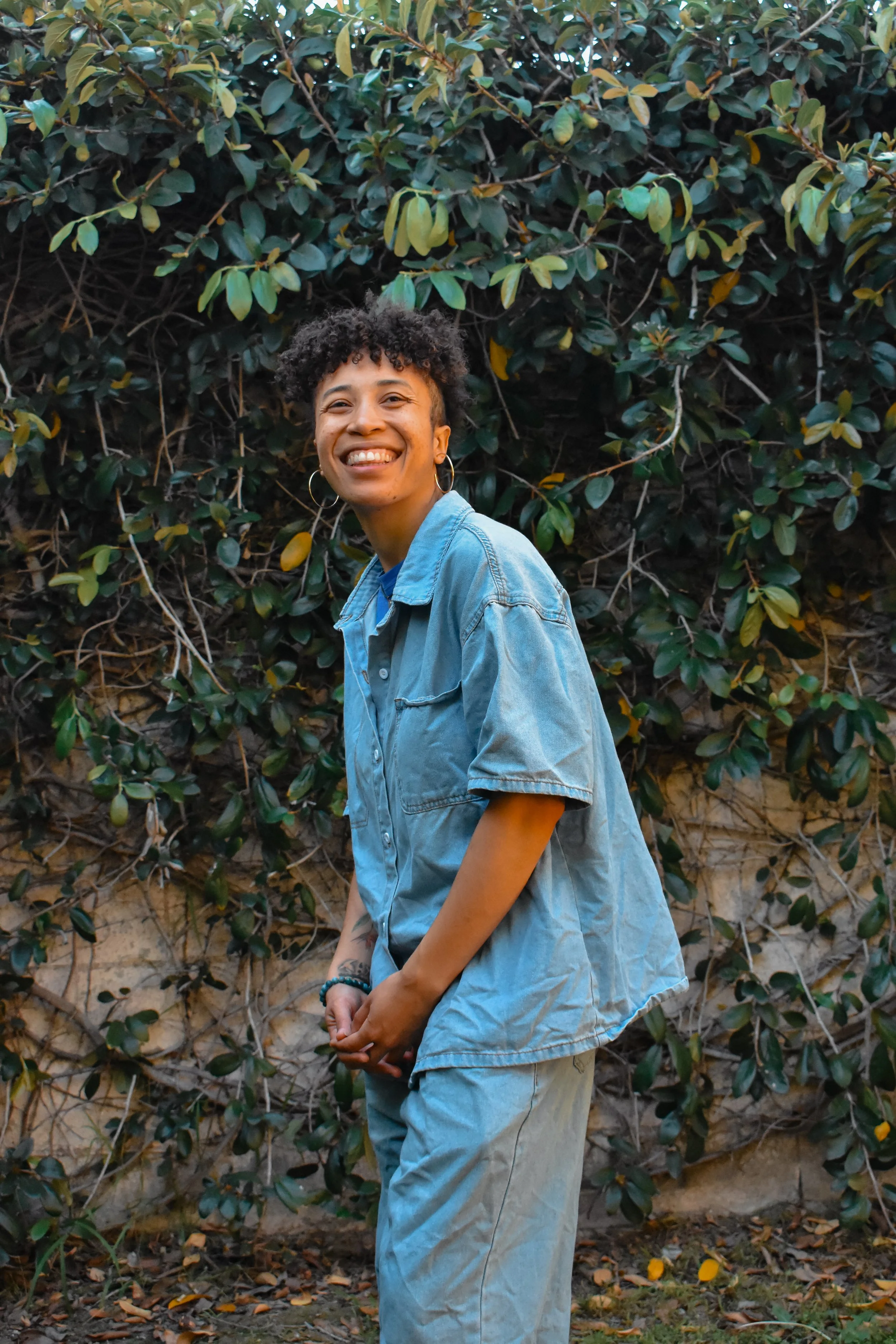 A person with short curly hair smiling and wearing a denim jacket stands in front of a green leafy bush.