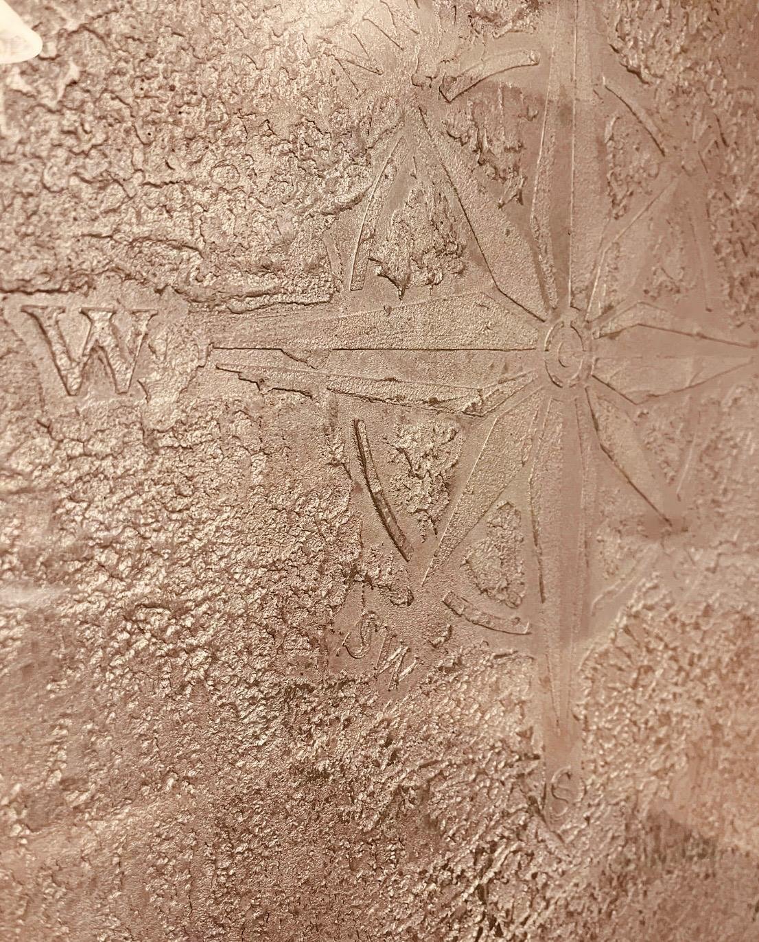 Close-up of a textured surface with a carved compass rose, the letter 'W' nearby, and some faint engraved text.