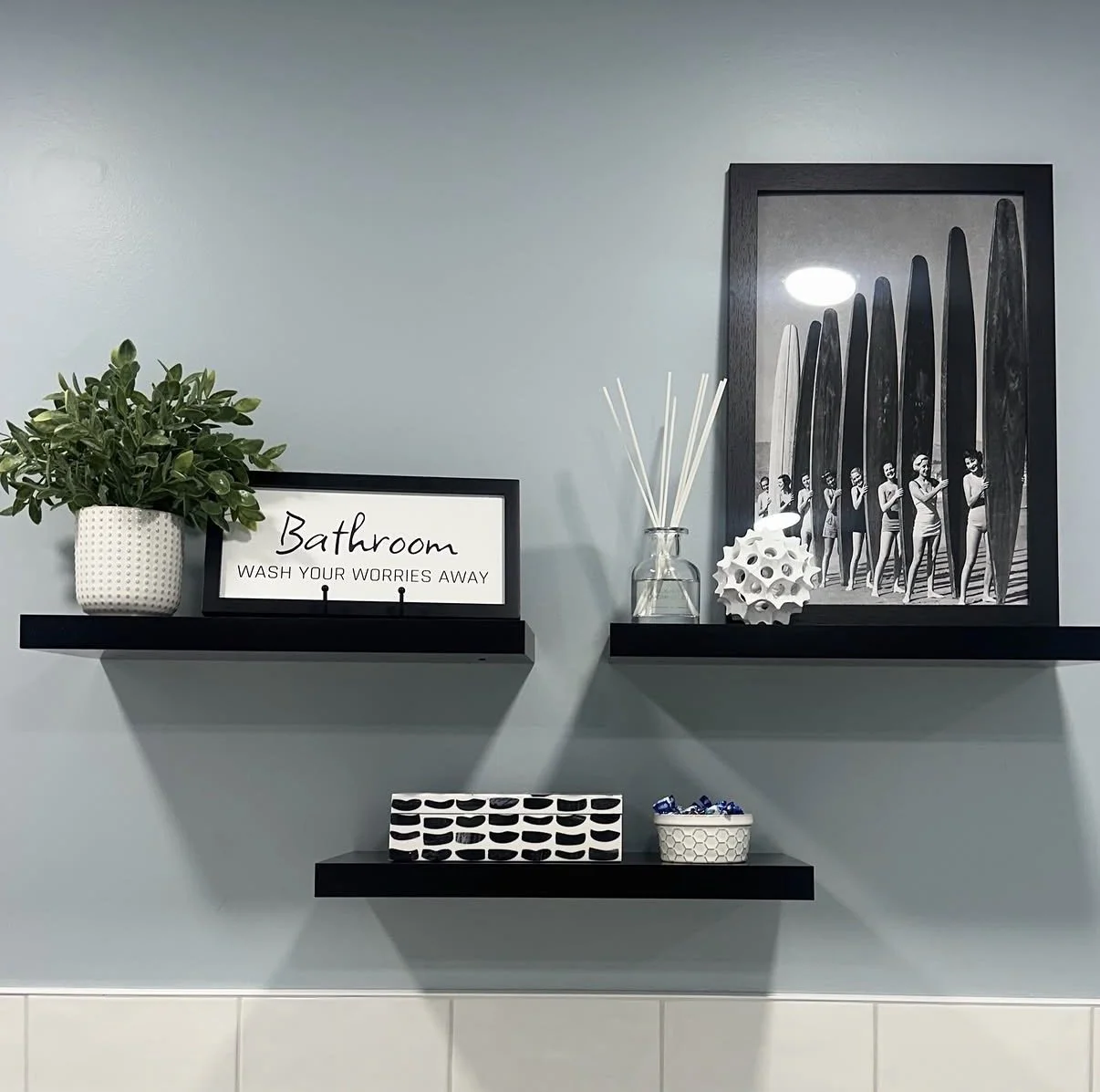 Decorative bathroom wall with black shelves holding a potted plant, a sign that says 'Bathroom Wash Your Worries Away', reed diffuser, sculptural object, and framed art of women with surfboards.