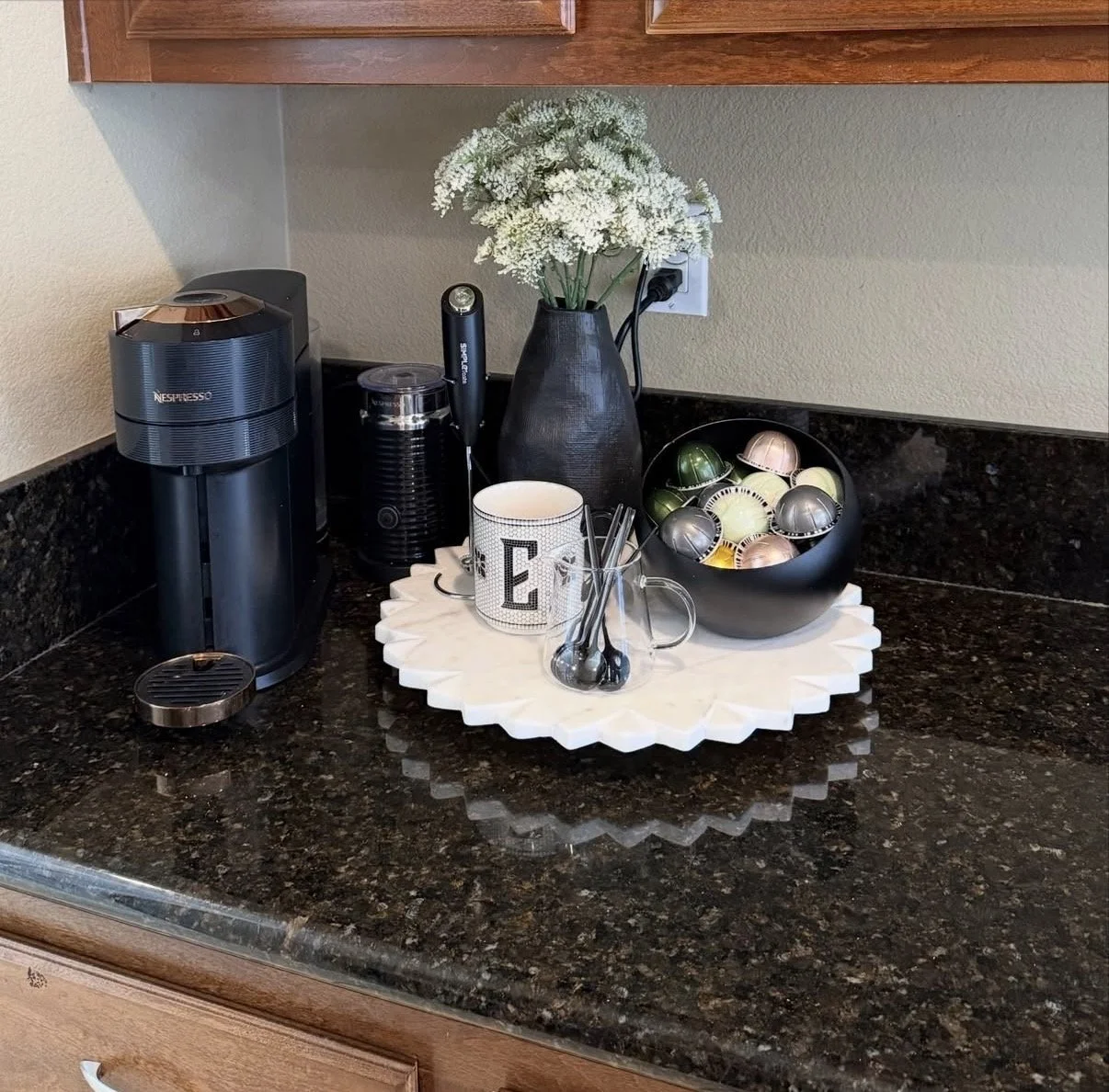 A kitchen countertop with a coffee machine, a mug, a small jar, a vase with white flowers, a bowl of coffee capsules, and a glass container with spoons on a white decorative tray.