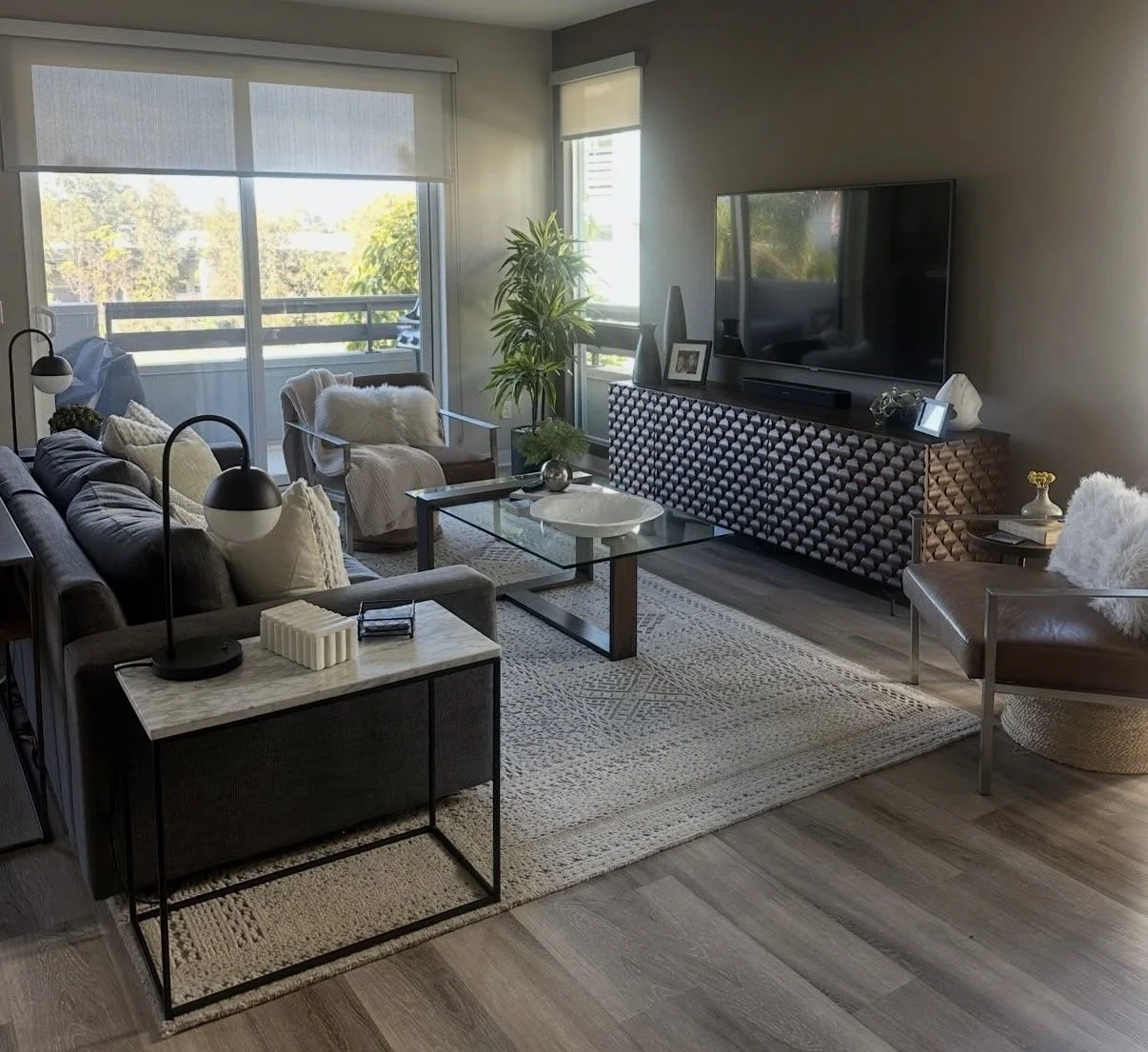 Living room with large windows, gray sofa, white armchair, glass coffee table, flat-screen TV on a decorative wooden console, various decorative items, potted plant, patterned area rug, and wooden flooring.