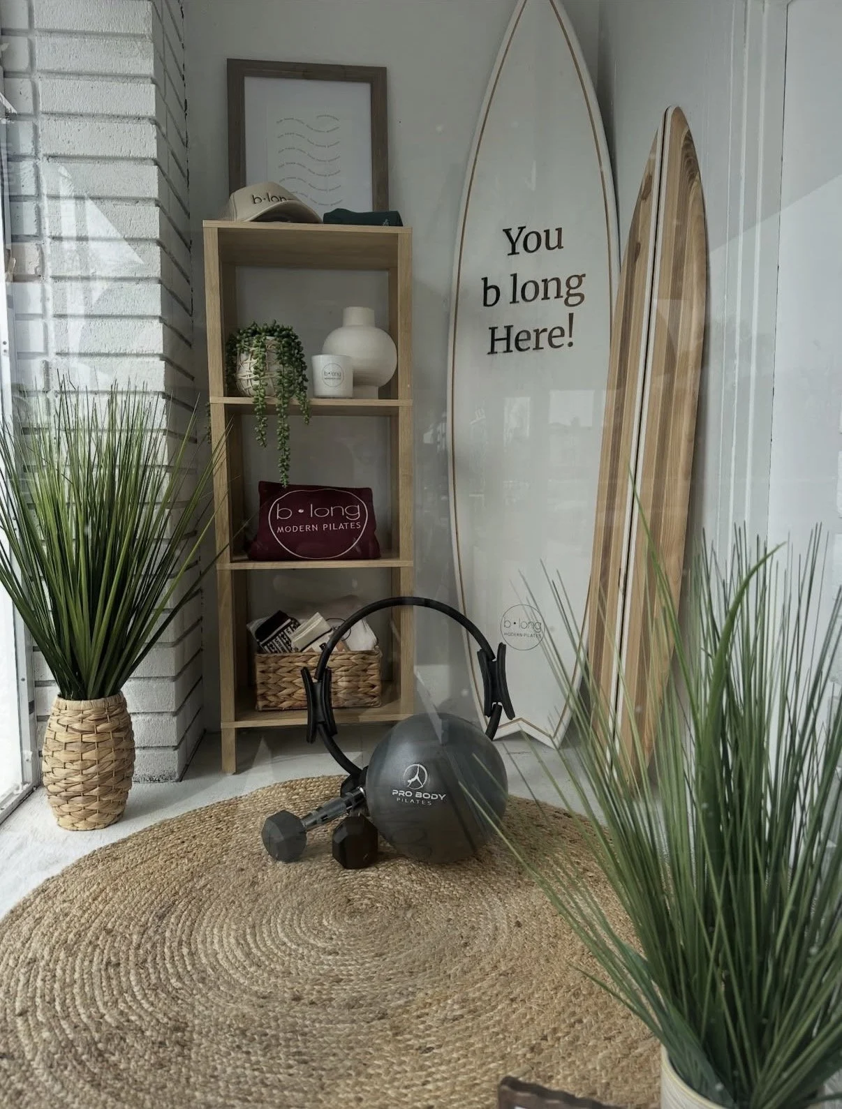 Interior of a fitness or wellness space with a wooden shelving unit holding plants, books, and decorative items. A large surfboard with the phrase "You b long Here!" leans against the wall. Fitness equipment, including a kettlebell and dumbbells, sit on a woven circular rug. Tall green plants in woven baskets are positioned on both sides of the room, enhancing the natural aesthetic.