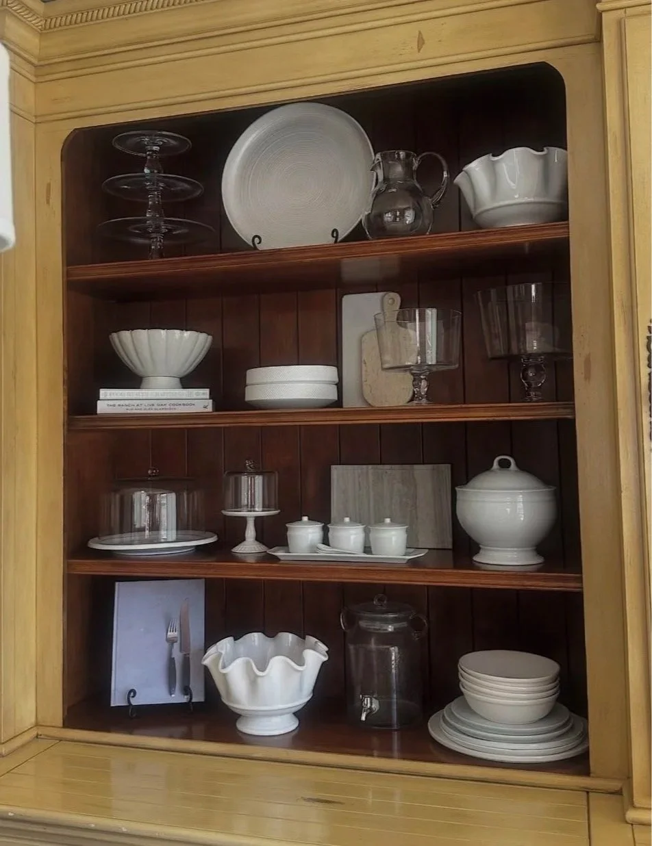 A wooden cabinet filled with white dishes, glassware, and serving bowls, including a large plate, bowl set, and various covered containers.