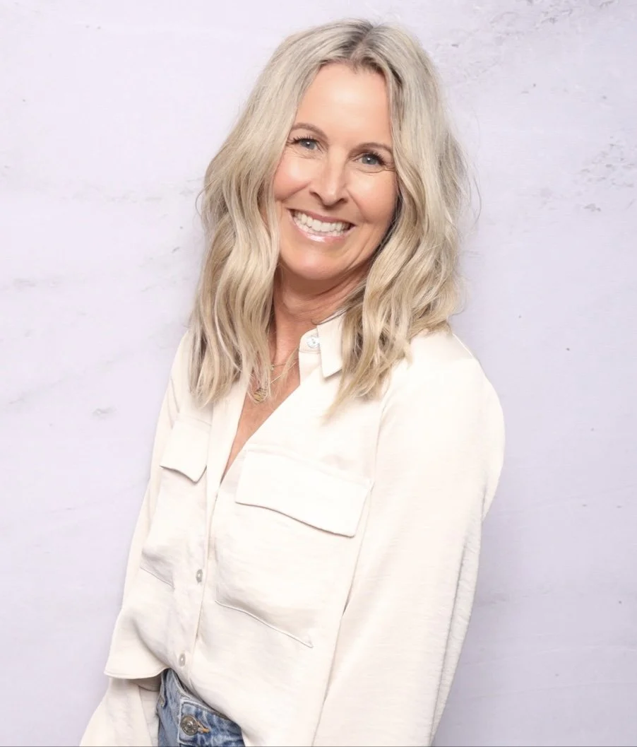 Smiling woman with blonde wavy hair wearing a white shirt and light blue jeans standing against a light gray background.