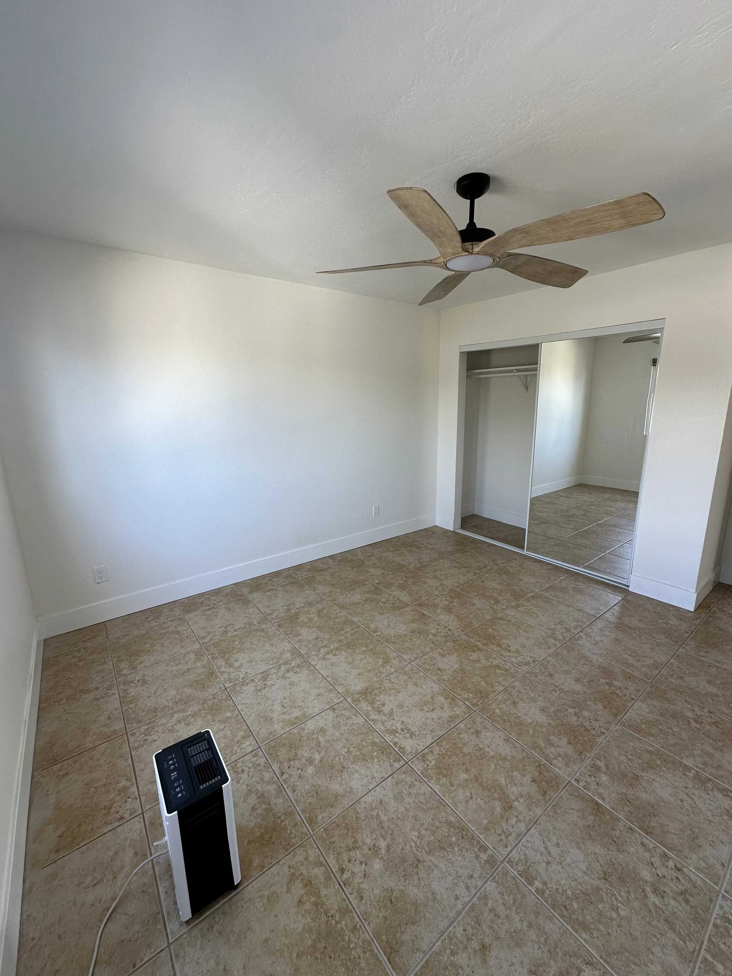 Empty room with tiled floor, white walls, a ceiling fan, a mirror closet, and a small air purifier.