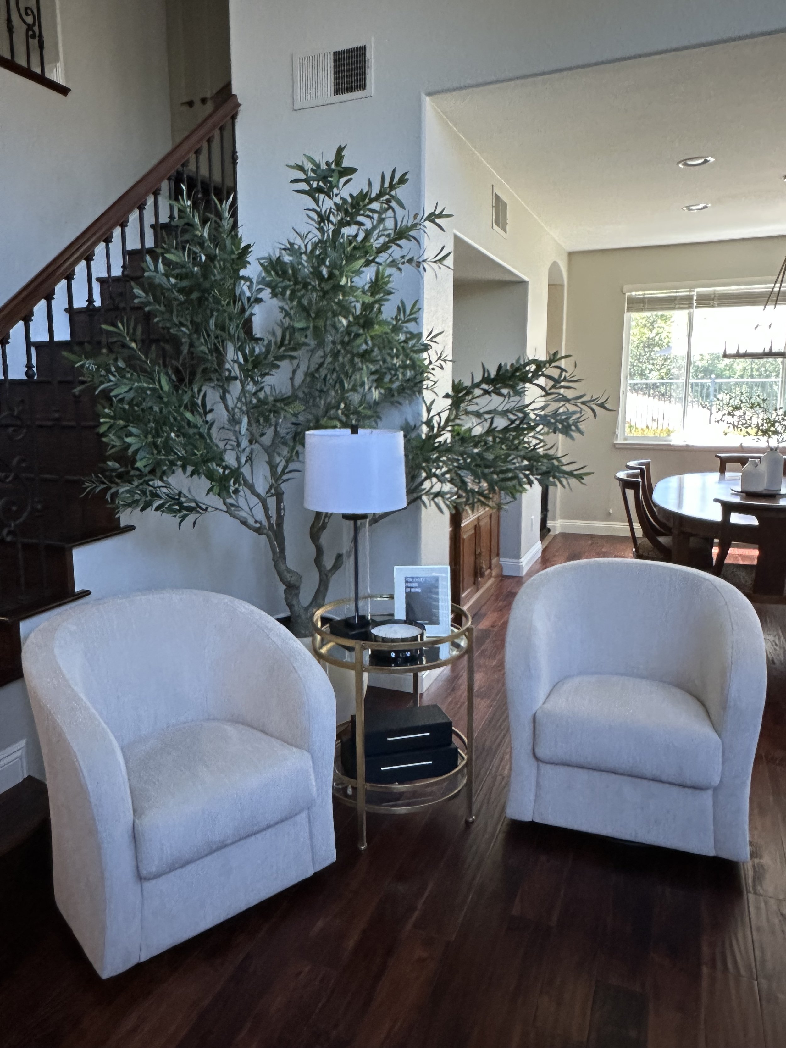 Living room with two white armchairs, a gold side table, a potted plant, and dining table near a large window.
