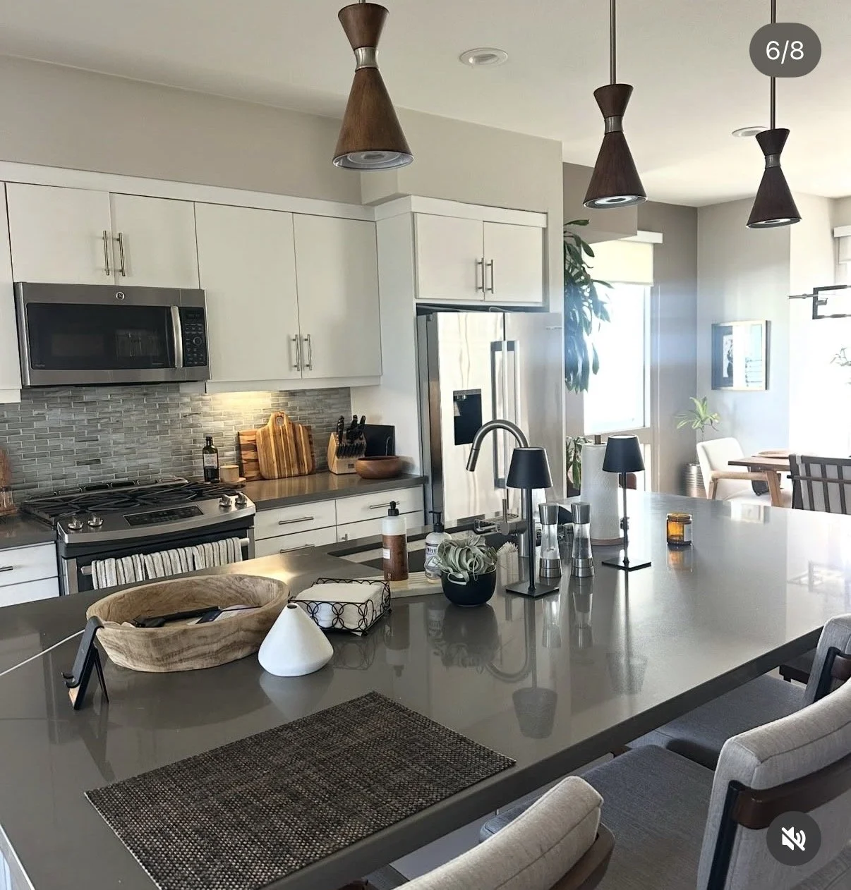 Modern kitchen with white cabinets, stainless steel appliances, and a center island with items including a wooden bowl, small plant, soap dispenser, and glass jars. Pendant lights hang above the island, and there is a dining area in the background with chairs near windows with blinds, natural light, and indoor plants.