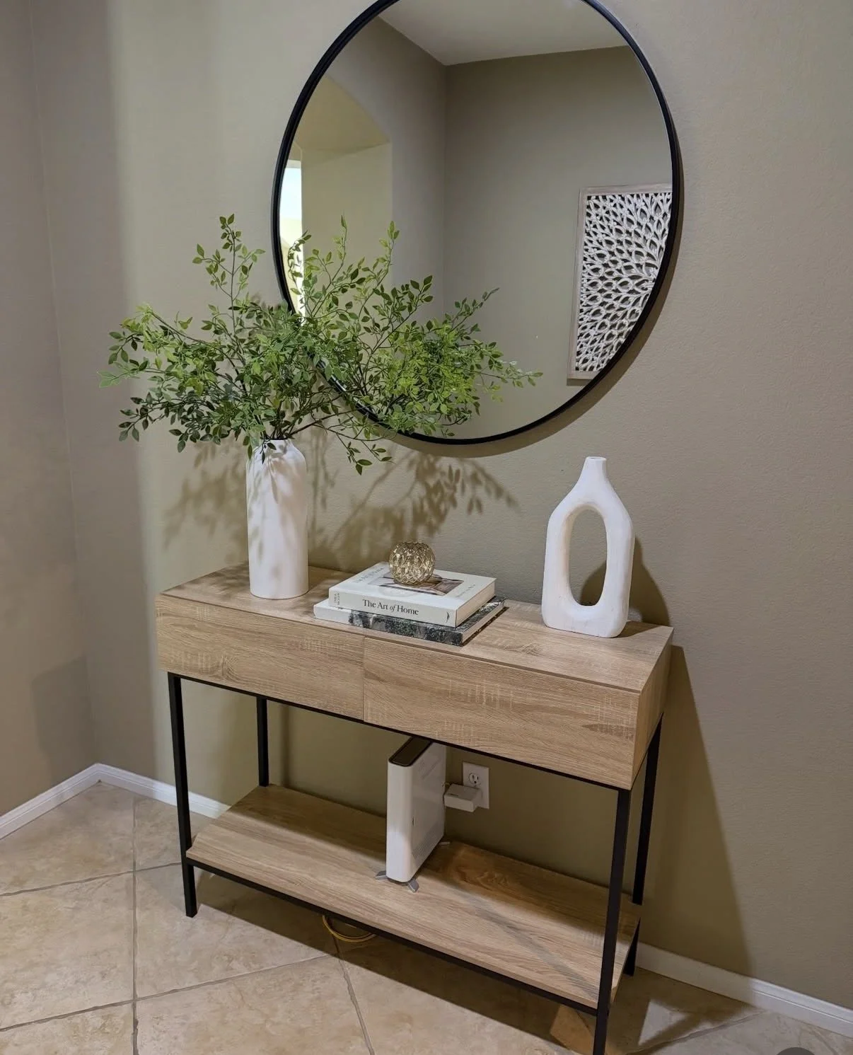 A wooden console table with black metal legs against a beige wall, decorated with a tall white vase holding green foliage, a white abstract sculpture, a small decorative sphere, and a stack of art and home books, with a large oval mirror above.