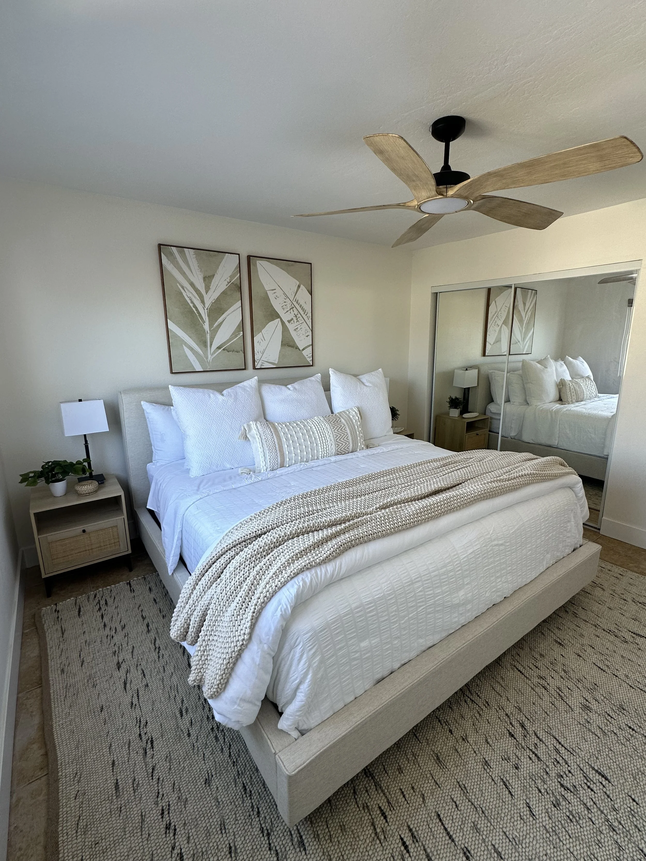 A bedroom with a white bed, beige knitted blanket, pillows, a nightstand with a plant and lamp, artwork of leaves on the wall, mirrored closet door, ceiling fan with wooden blades, and a patterned area rug.