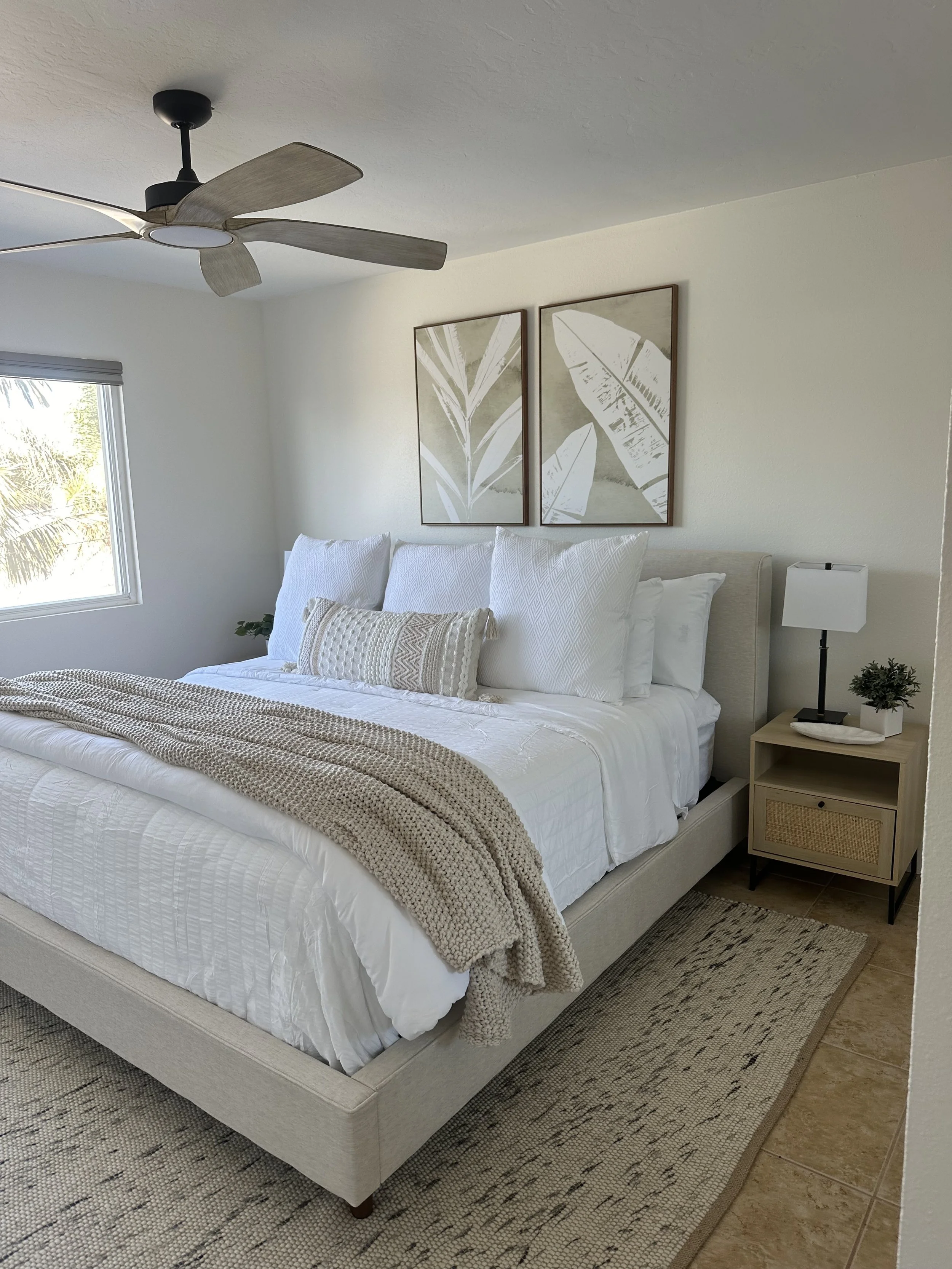 A bedroom with a bed dressed in white linens, a knitted beige blanket, and multiple white pillows. A bedside table holds a white lamp and a small plant. Two framed botanical prints hang above the bed. There is a window with a view of palm trees and a ceiling fan with wooden blades.