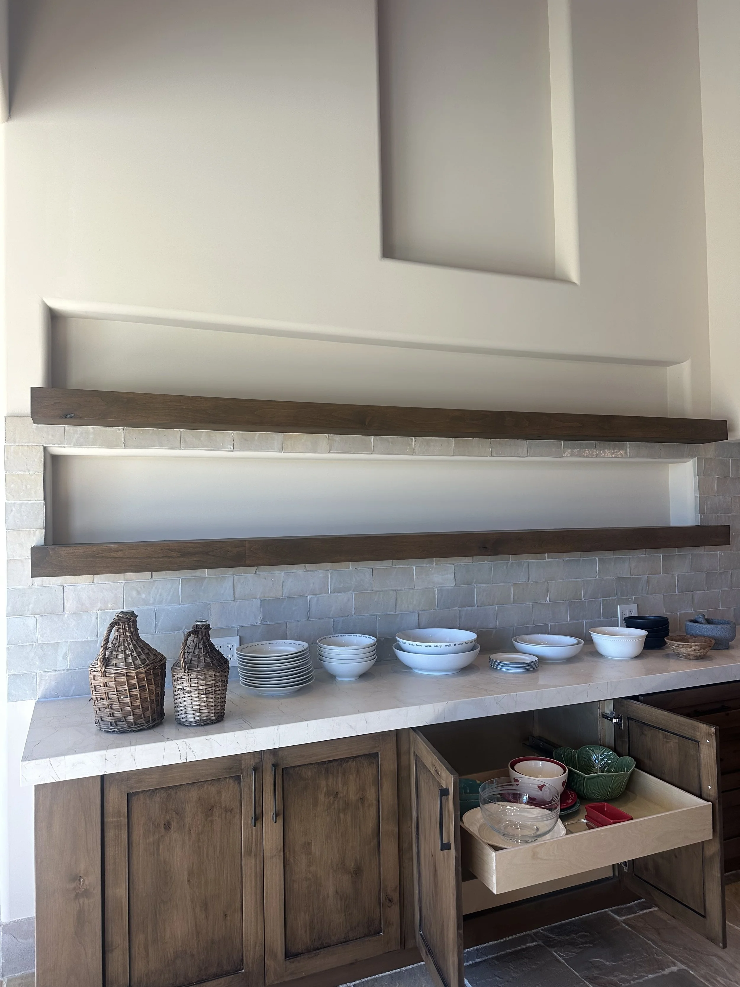 Kitchen counter with stacked bowls, woven baskets, and dishes, beneath wooden cabinets and open shelves.