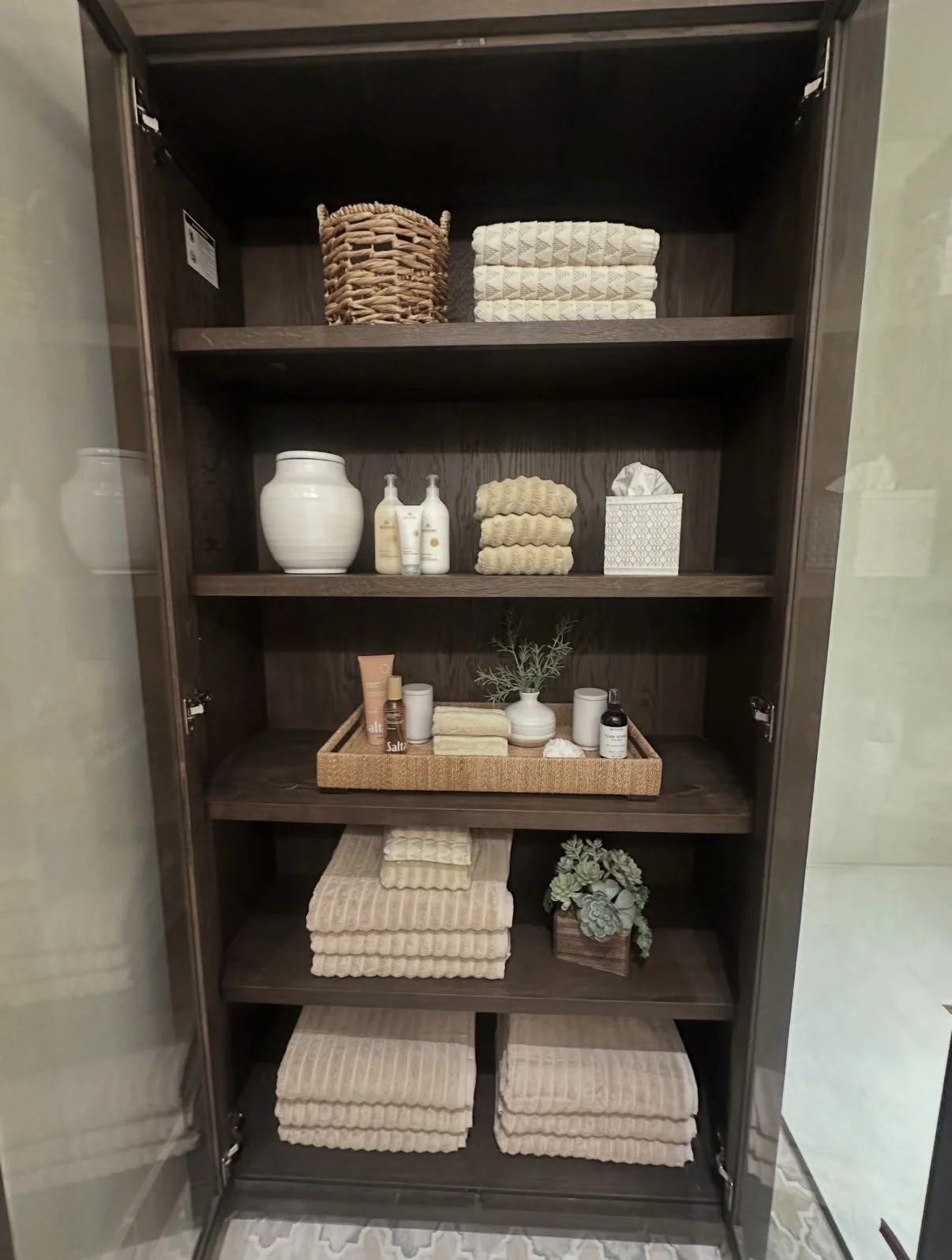 Open dark wood cabinet with five shelves containing neatly folded towels, baskets, skincare products, a white ceramic vase, a small plant, and a box of tissues.