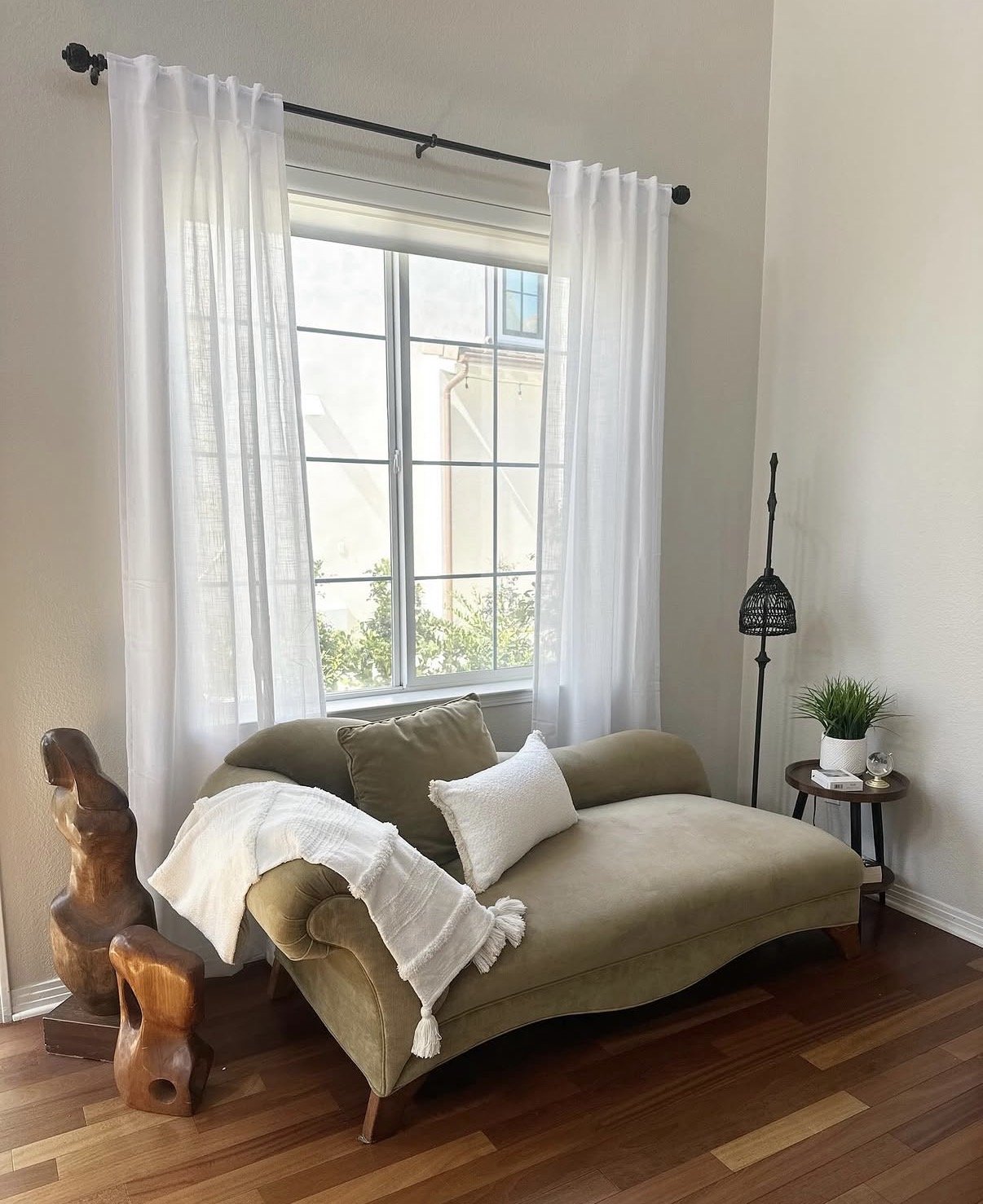 A cozy living room corner with a green vintage sofa, white throw blanket, and pillows near a window with sheer white curtains, a small side table with a potted plant, a candle, and a modern floor lamp.