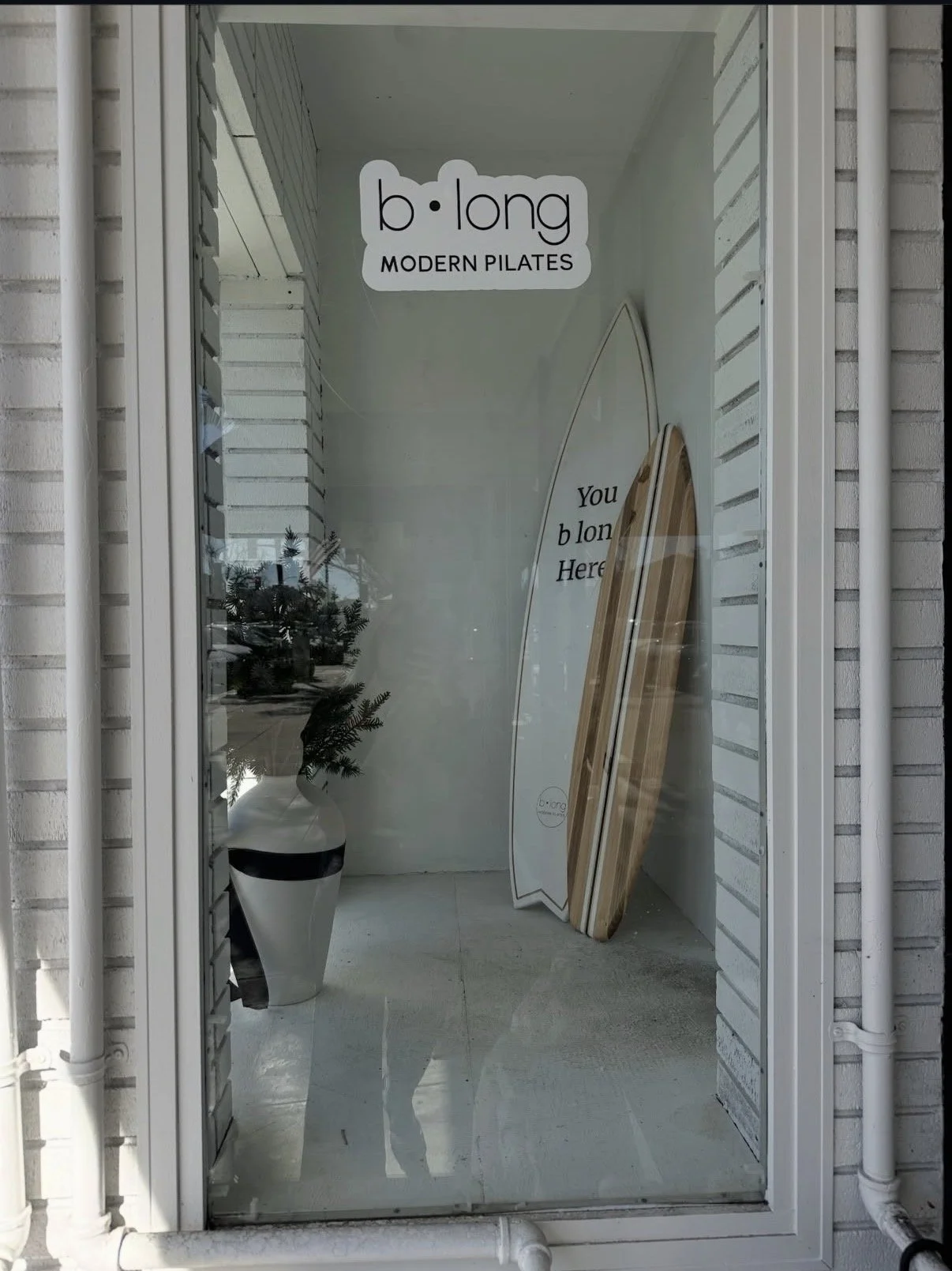 Storefront window with a sign that reads 'b•long MODERN PILATES' and a surfboard with the words 'You b long Here' written on it, with a decorative plant inside.