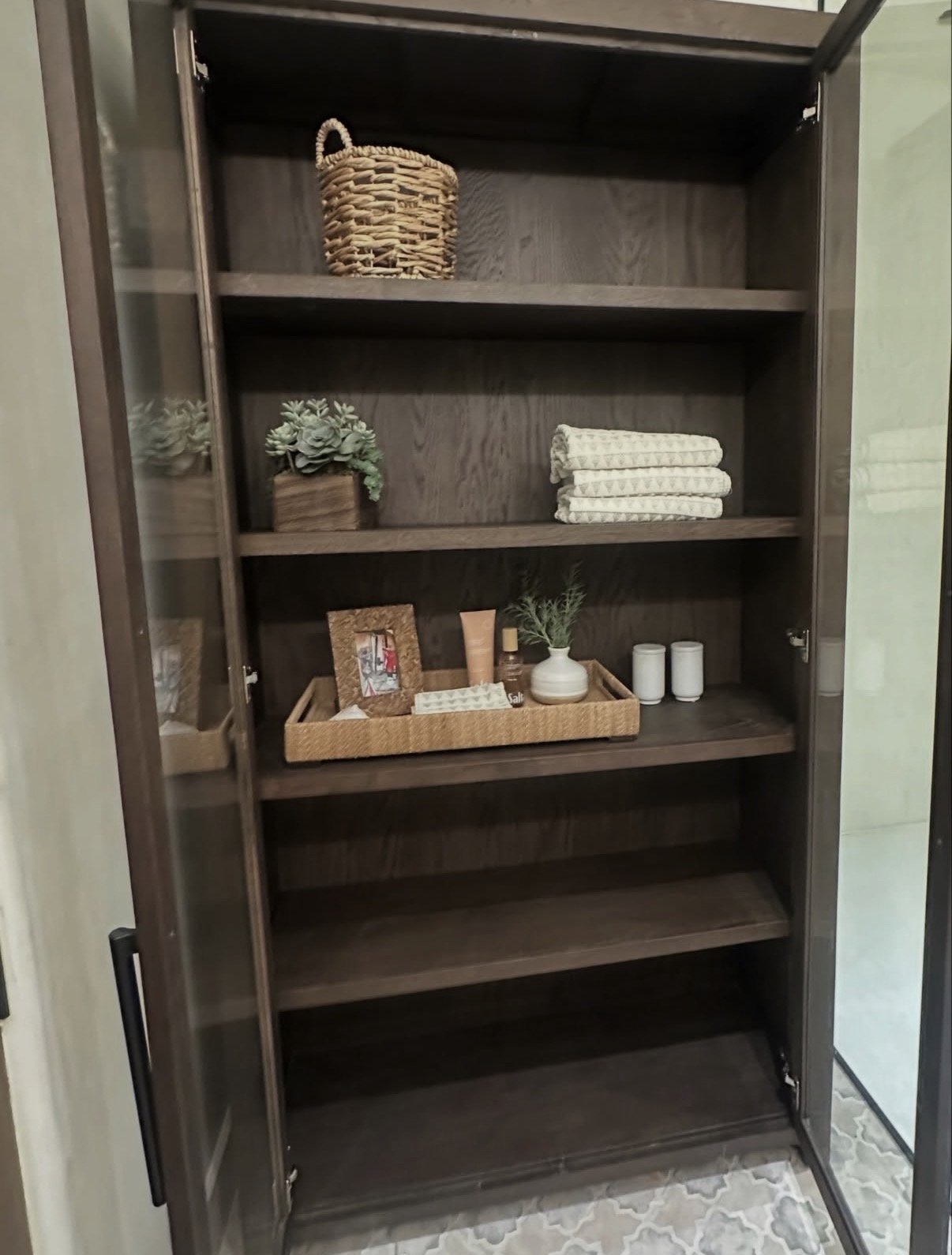A dark wooden cabinet with glass doors on either side, containing shelves with decorative items such as a wicker basket, small plant, folded linens, picture frame, lotion, and candles.