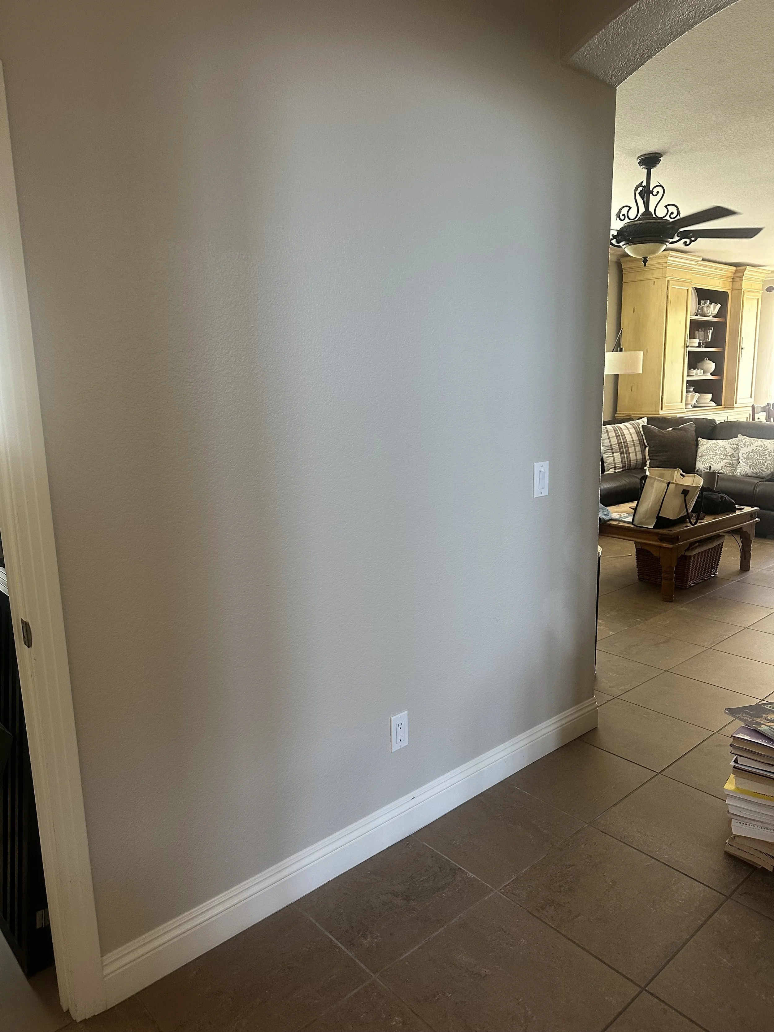 Interior of a house with a beige wall, dark brown tiled floor, white baseboard, and a view into a living room area with a ceiling fan, wooden cabinet, sofa with pillows, and various items on a coffee table.