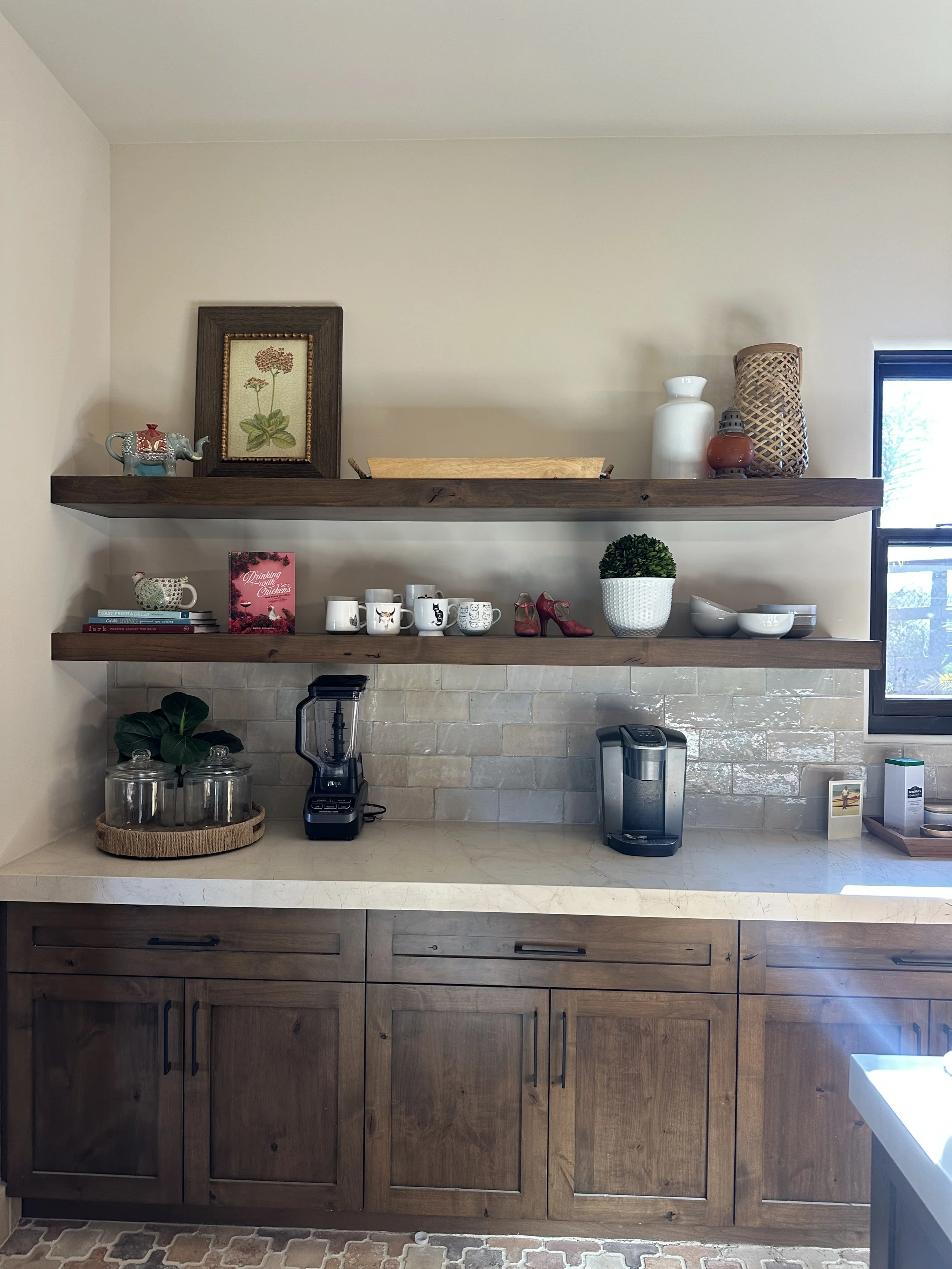 Kitchen countertop with three wooden floating shelves holding various decorative items, mugs, a potted plant, and kitchen appliances, with a window on the right side.