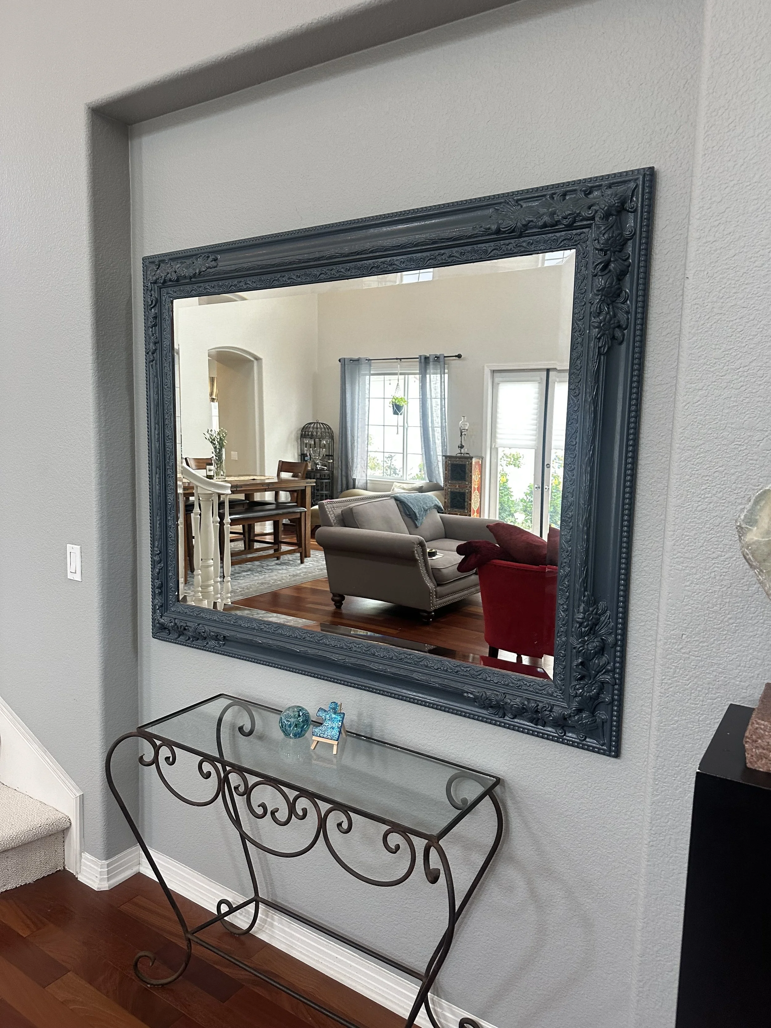 Reflection of a living room with a beige sofa, red armchair, dining table with chairs, and various decor items, seen in a large ornate blue-gray mirror on a white wall.