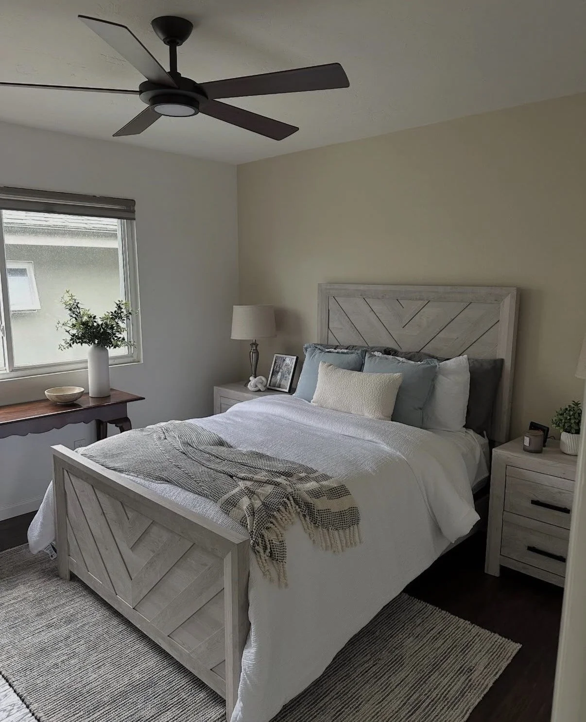 A bedroom with a white wooden bed, blue and white pillows, a blanket, and a beige throw. There is a wooden nightstand with a lamp, picture frame, and a small plant. A window with a vase and green plant on a wooden table is visible, along with a ceiling fan and a rug on the dark wood floor.