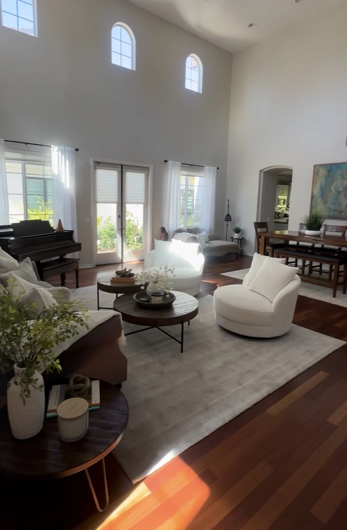 A spacious living room with high ceilings, multiple windows letting in natural light, and modern white furniture, including a sofa and armchair, arranged around a round coffee table with decorative items. The room features wooden flooring and a large painting on the wall, with a dining table and chairs against one wall, and a piano near the windows.