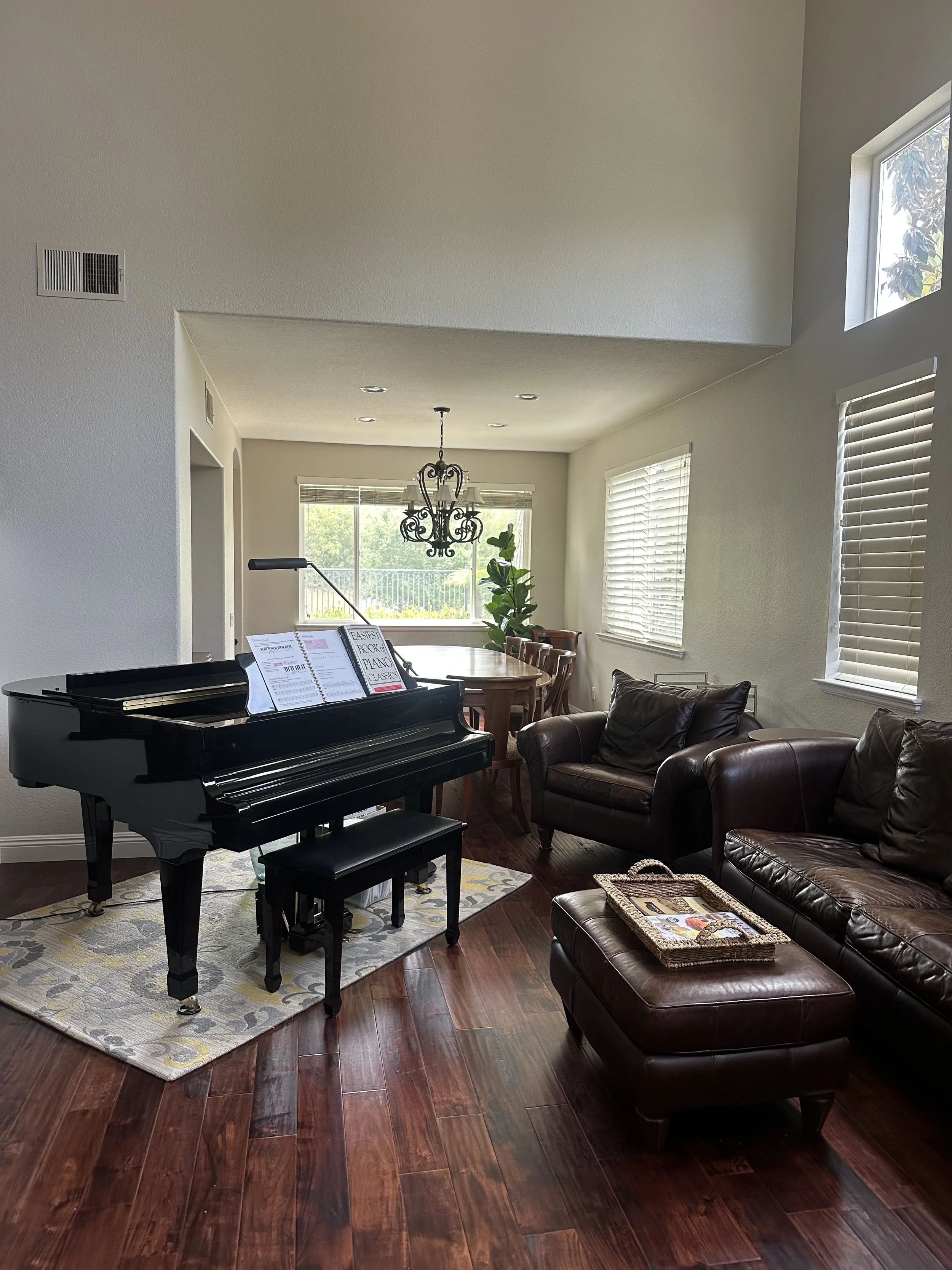 Living room with a piano, leather sofas, wooden dining table with chairs, chandelier, and large windows with blinds.
