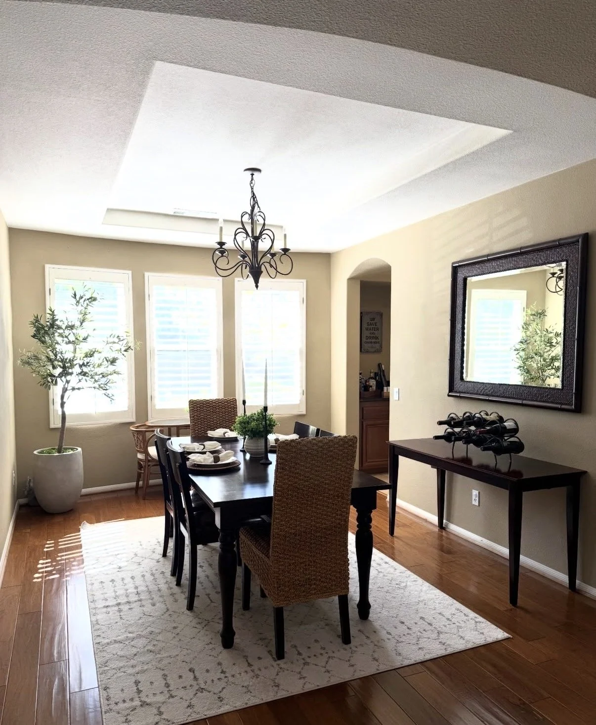 A dining room with a dark wooden table set for a meal, six chairs, a chandelier, three windows with blinds, a large potted plant, a mirror on the wall, and a side table with wine bottles.
