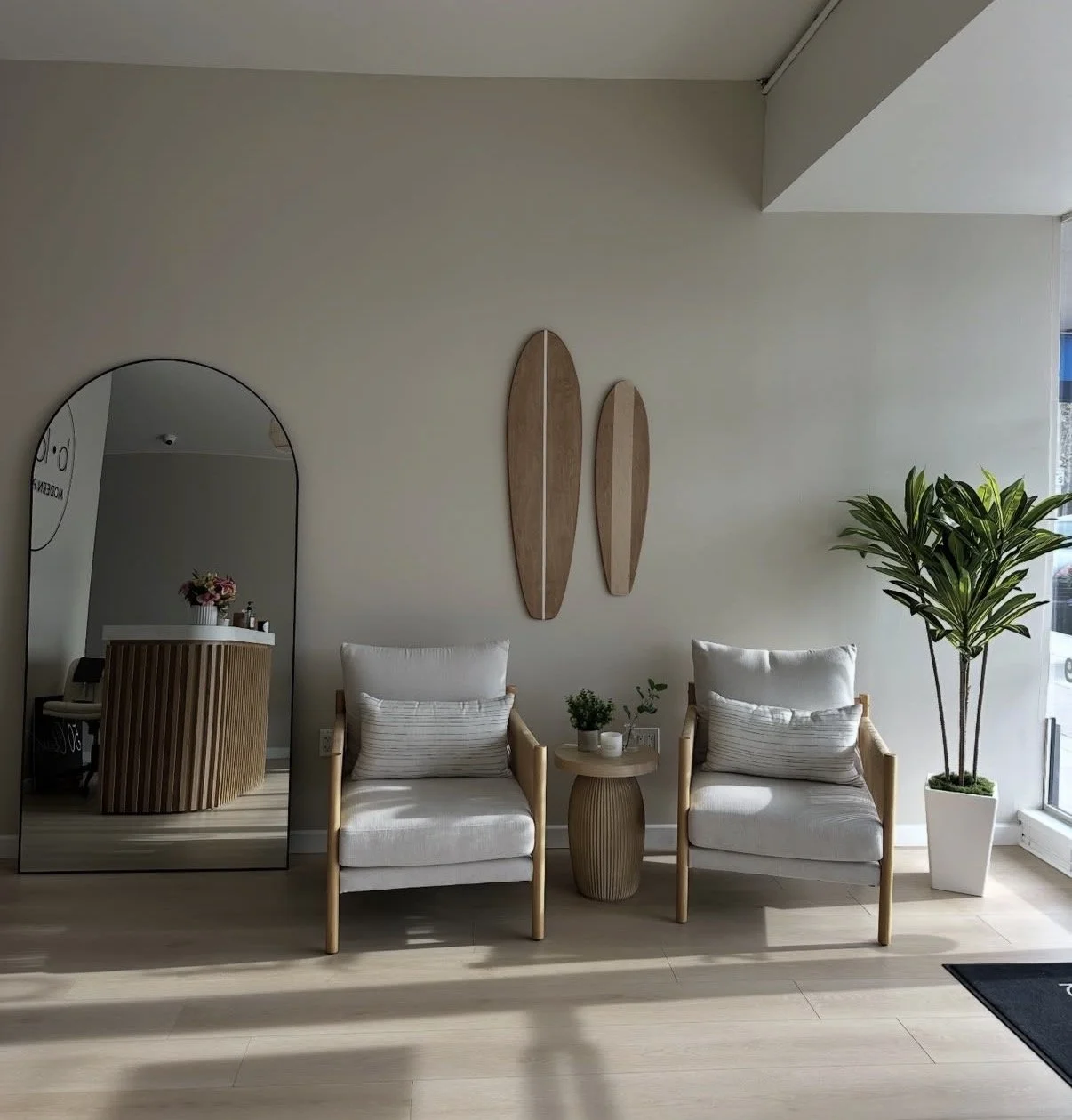 Office waiting area with two white cushioned chairs, a small round wooden table with potted plants, a large potted plant in a white planter, a mirror reflecting another area, and wall art with two wooden oval shapes.