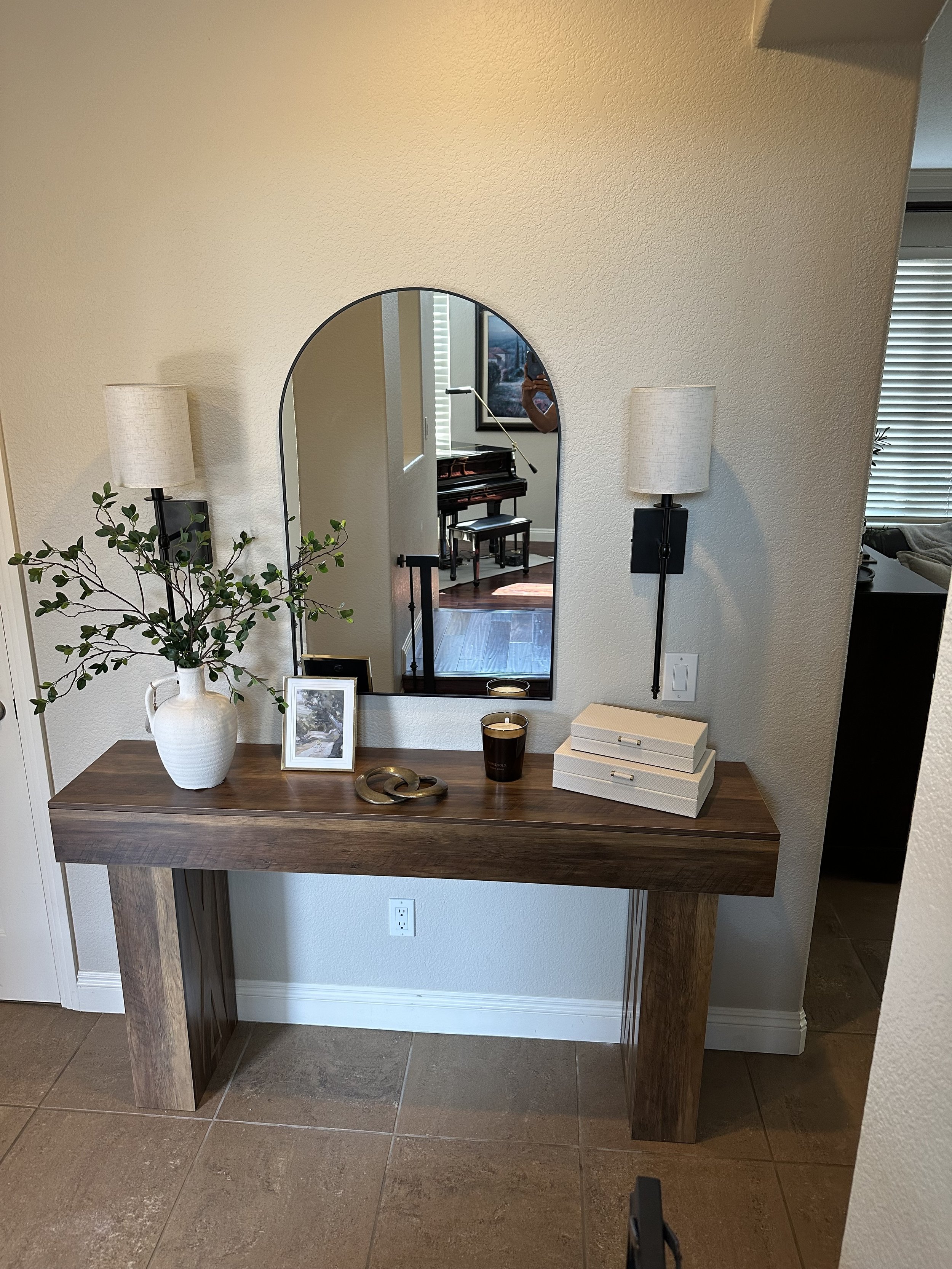 A wooden console table with a white vase containing green leafy branches, a framed black and white photo, two lit candles, and stacked boxes. A large mirror with an arched top hangs above the table, reflecting a grand piano and a person taking the photo.