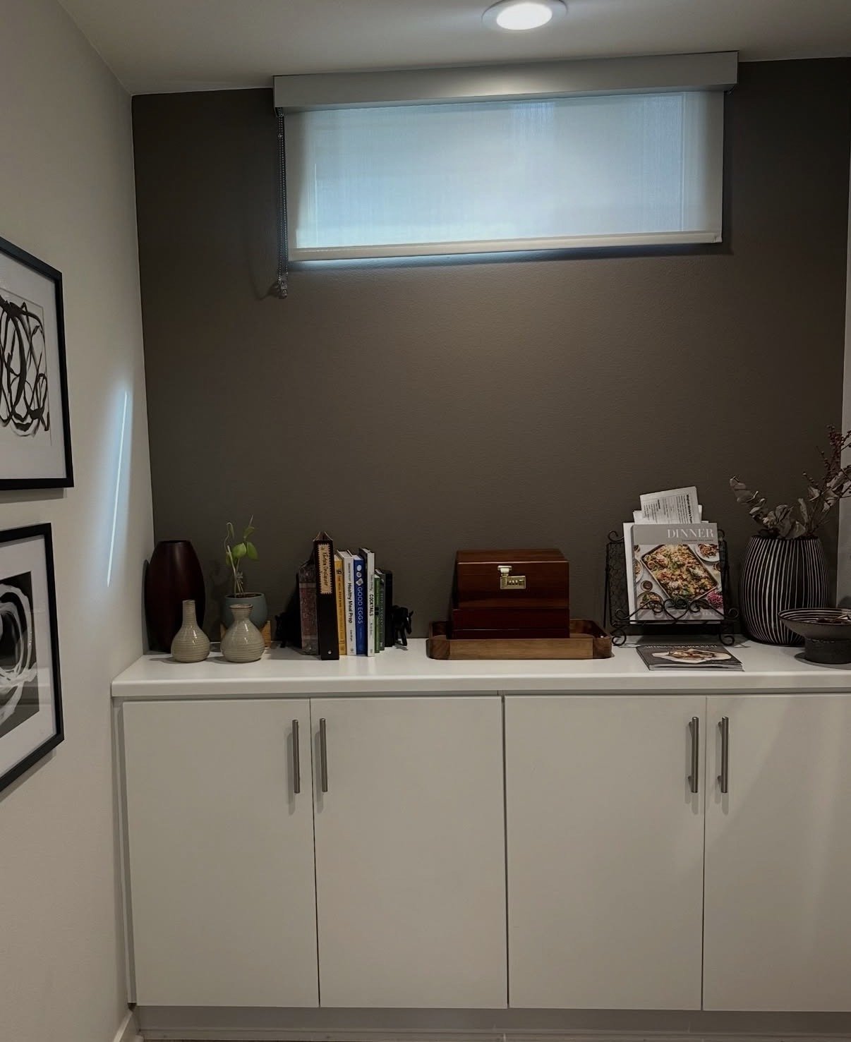 A white cabinet with decorative vases, books, a wooden box, and a magazine stand holding a dinner recipe book, topped with a planter with dried branches, in a room with a high window and dark gray wall.