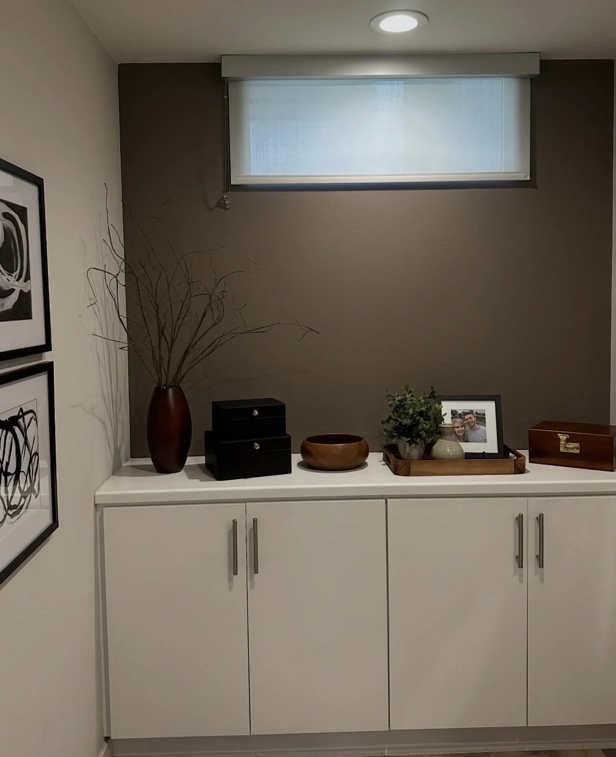 Decorative sideboard with a tall brown vase of branches, small black storage boxes, a wooden bowl, a tray with a potted plant and a picture frame, and a wooden jewelry box, with artwork on the wall and a window above.