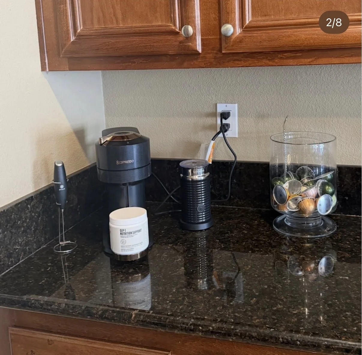 Kitchen countertop with a coffee machine, a container of nutritional support, a milk frother, a jar filled with various K-Cup pods, and a pod whisk nearby, with wooden cabinets above and an electrical outlet on the wall.