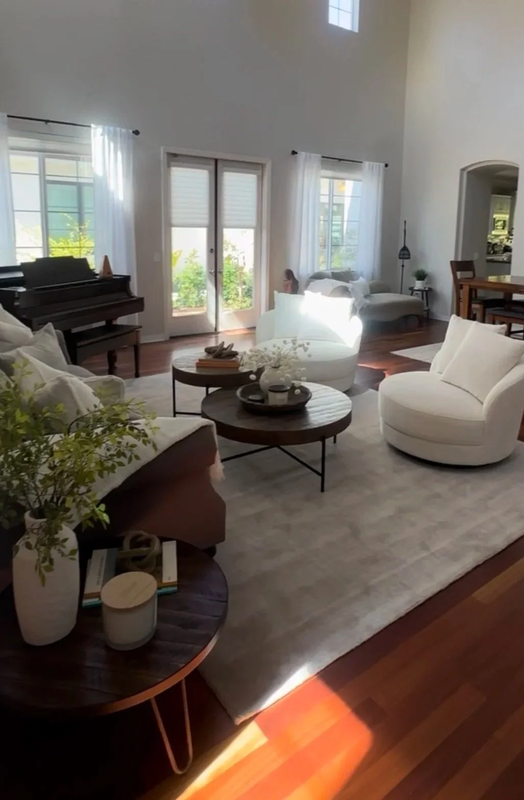 Bright living room with white sofas, a round coffee table, and large windows with sheer curtains, featuring hardwood floors and a white area rug.