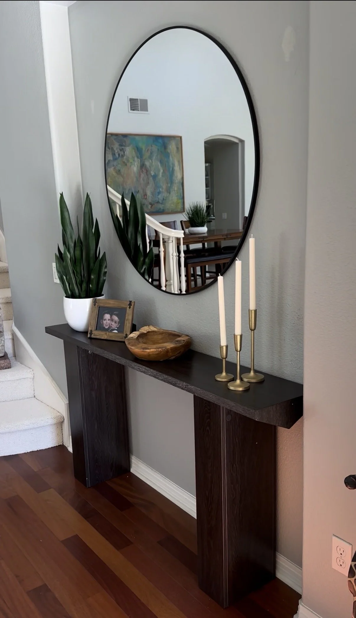Home interior featuring a rectangular dark wood console table with decorative items including white candles, a wooden bowl, a framed photo, and a potted plant. Above the table is a large oval mirror on a gray wall, and part of a staircase with white carpeted steps is visible to the left.