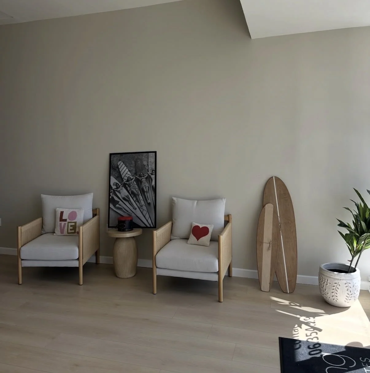 Living room corner with two white armchairs, pillows with love-themed designs, a small wooden side table, and a skateboard leaning against the wall near a potted plant.