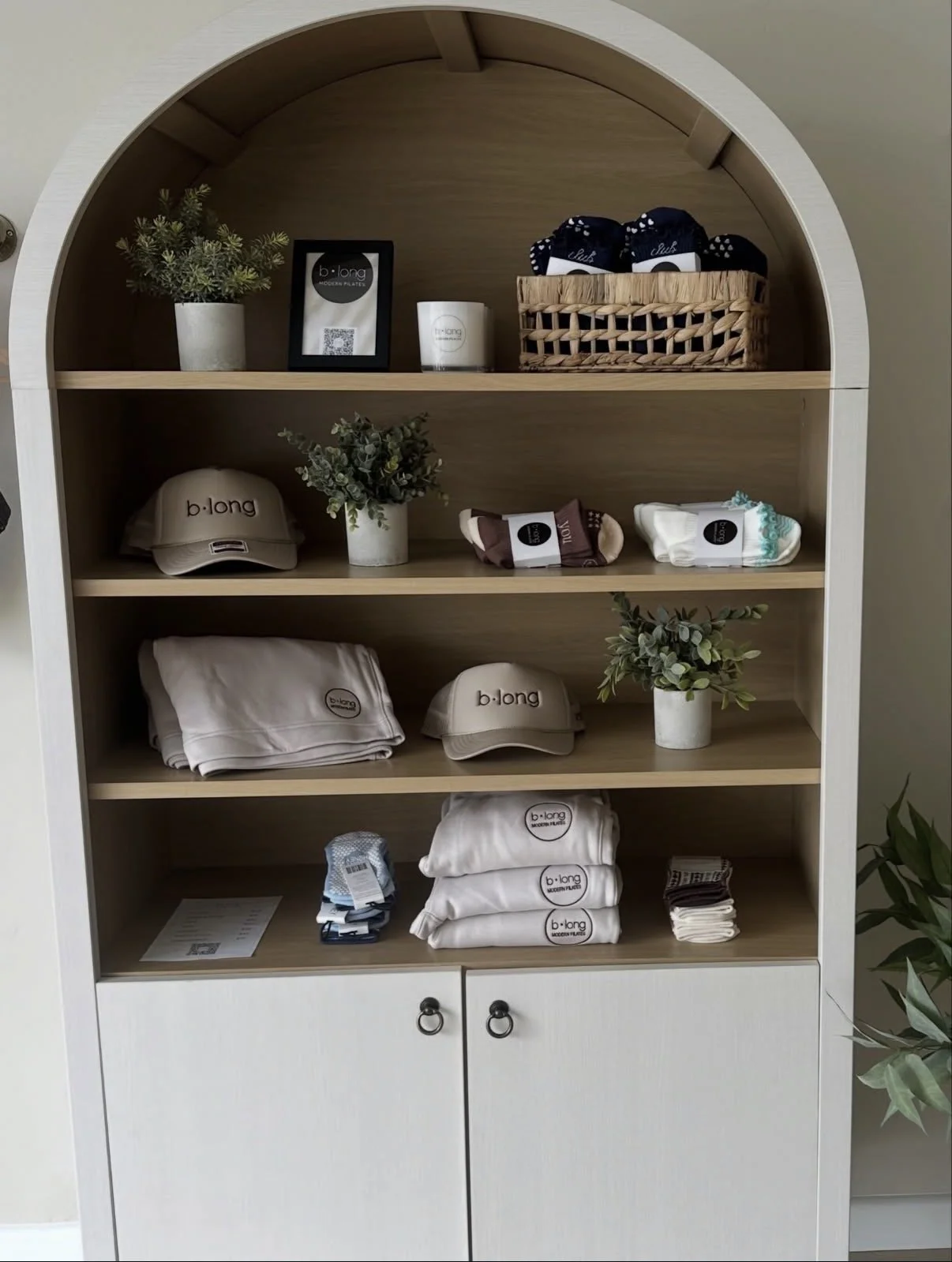 A white wooden shelf with four open shelves and a closed cabinet at the bottom, displaying clothing and accessories. The top shelf has a potted plant, a framed picture, a candle, and a basket of socks. The second shelf has a beige hat, a potted plant, and rolled socks. The third shelf has folded clothing, a hat, and a potted plant. The bottom shelf has folded clothing, a hat, and a small stack of socks.