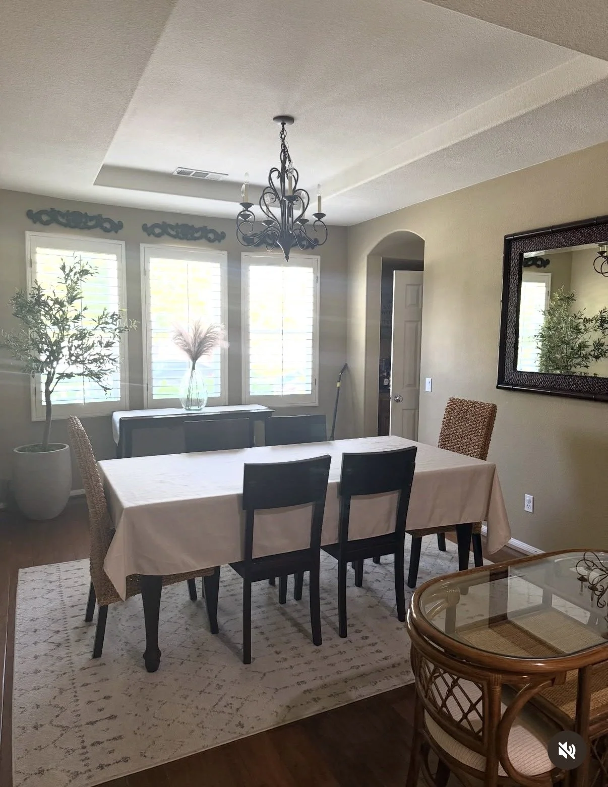 A dining room with a table covered in a white tablecloth, surrounded by black and wicker chairs, a chandelier hanging from the ceiling, and three windows with white shutters allowing sunlight to brighten the room. There are two potted plants, a mirror on the right wall, and a glass-topped wicker side table in the foreground.