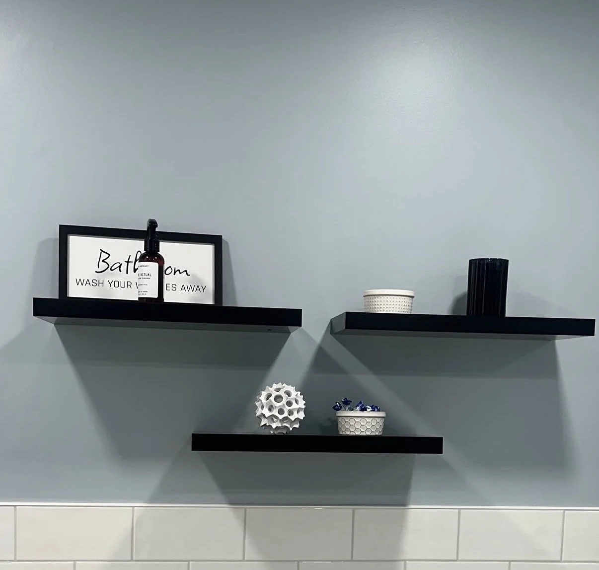Three black floating shelves on a light blue wall with decorative items including a framed sign, bottles, a bowl, and geometric sculptures.
